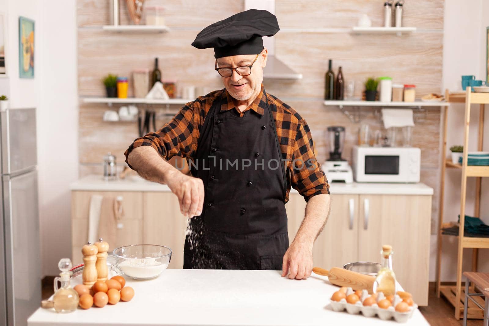 Senior chef preparing homemade bread sprinkling wheat flour on kitchen table. Retired senior chef with bonete and apron, in kitchen uniform sprinkling sieving sifting ingredients by hand.