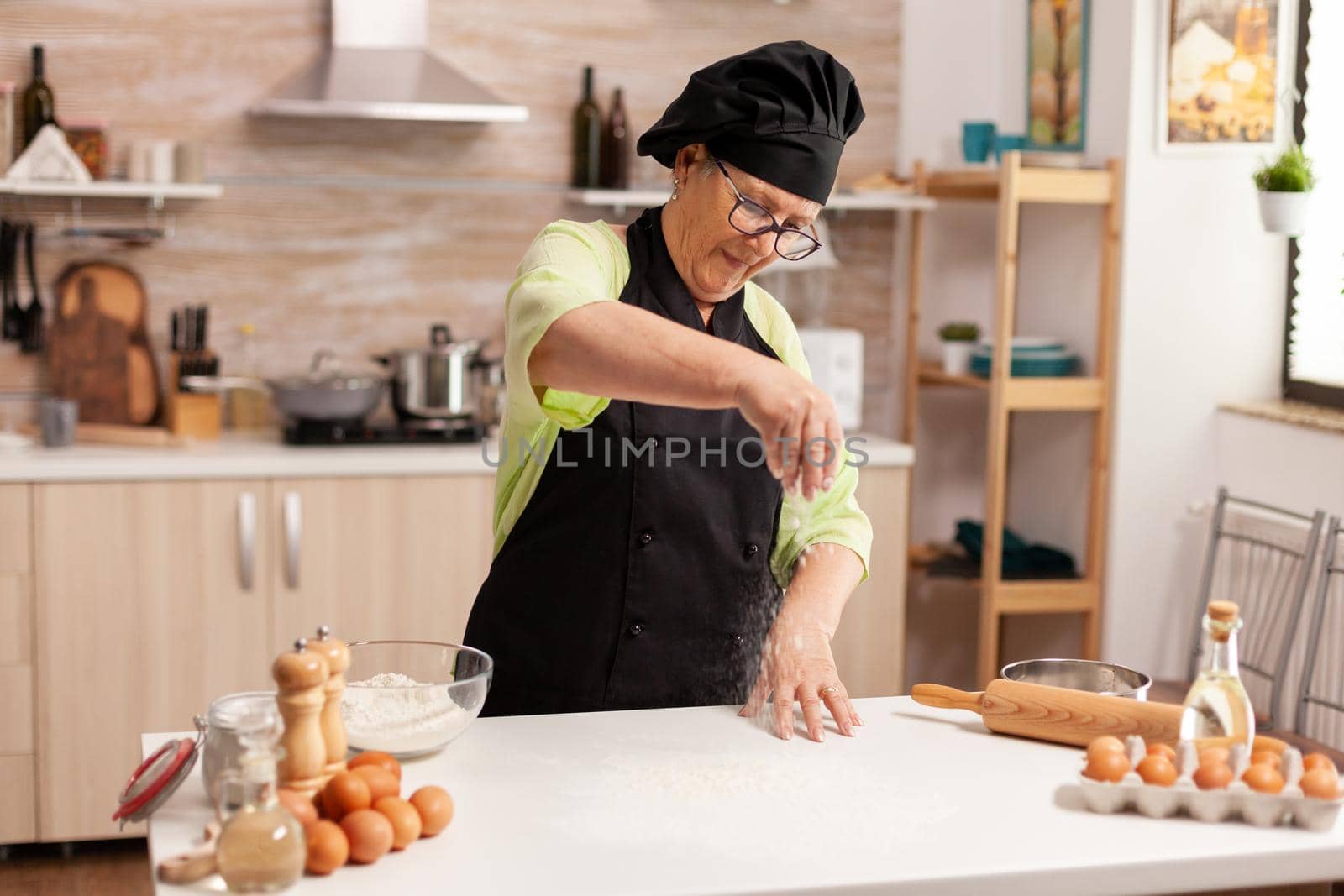 Elderly woman using flour to make delicious cookies on home kitchen table spreading flour. Happy elderly chef with uniform sprinkling, sieving sifting raw ingredients by hand.