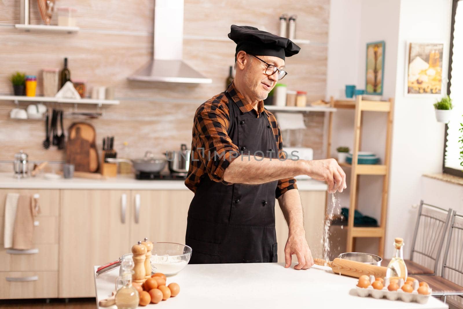 Retired senior baker wearing apron and bonete using ingredients for homemade pizza. Chef with in kitchen uniform sprinkling sieving sifting ingredients by hand.