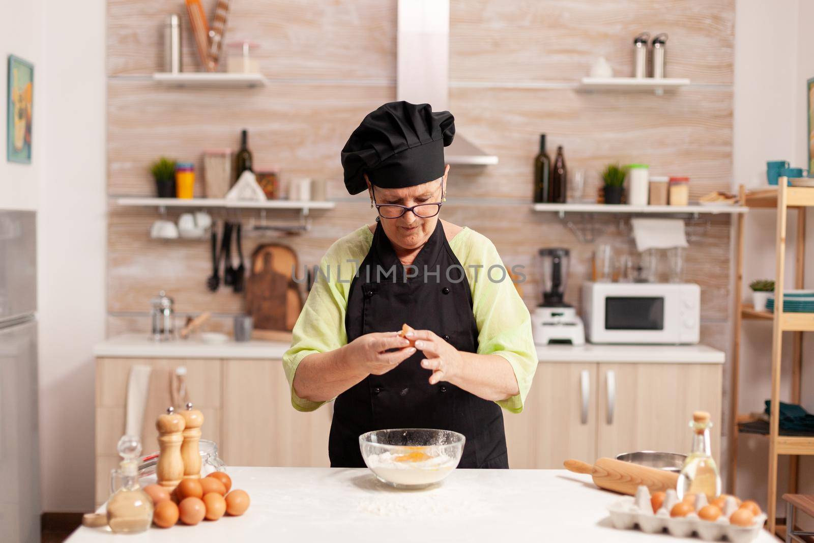 Chef hands with flour and eggs in preparation process for baking. Elderly pastry chef cracking egg on glass bowl for cake recipe in kitchen, mixing by hand, kneading.