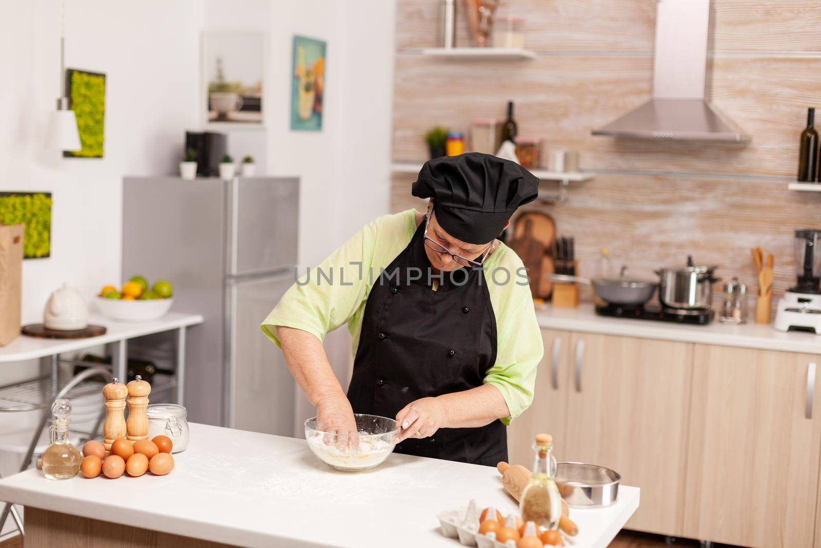 Woman mixing eggs with flour to make dough after traditional recipe on kitchen table. Retired elderly chef with uniform sprinkling, sieving sifting raw ingredients and mixing .