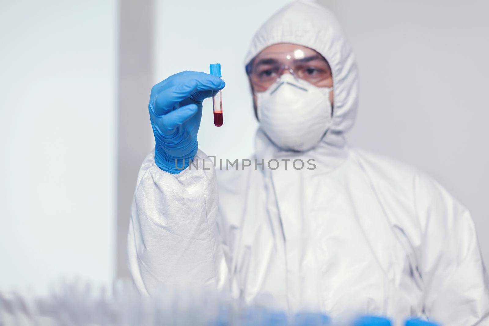 Man in coverall suit in microbiology laboratory holding test tube with blood infected with coronavirus. Doctor working with various bacteria and tissue, pharmaceutical research for antibiotics against covid19.