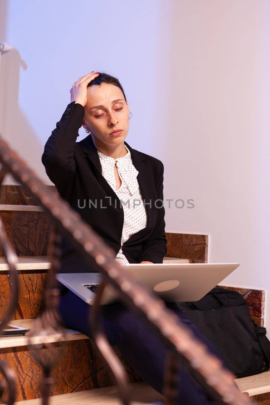 Stressed overworked businesswoman doing overtime for project deadline at late hours. Serious entrepreneur working on job project sitting on staircase of business building at night hours for job.