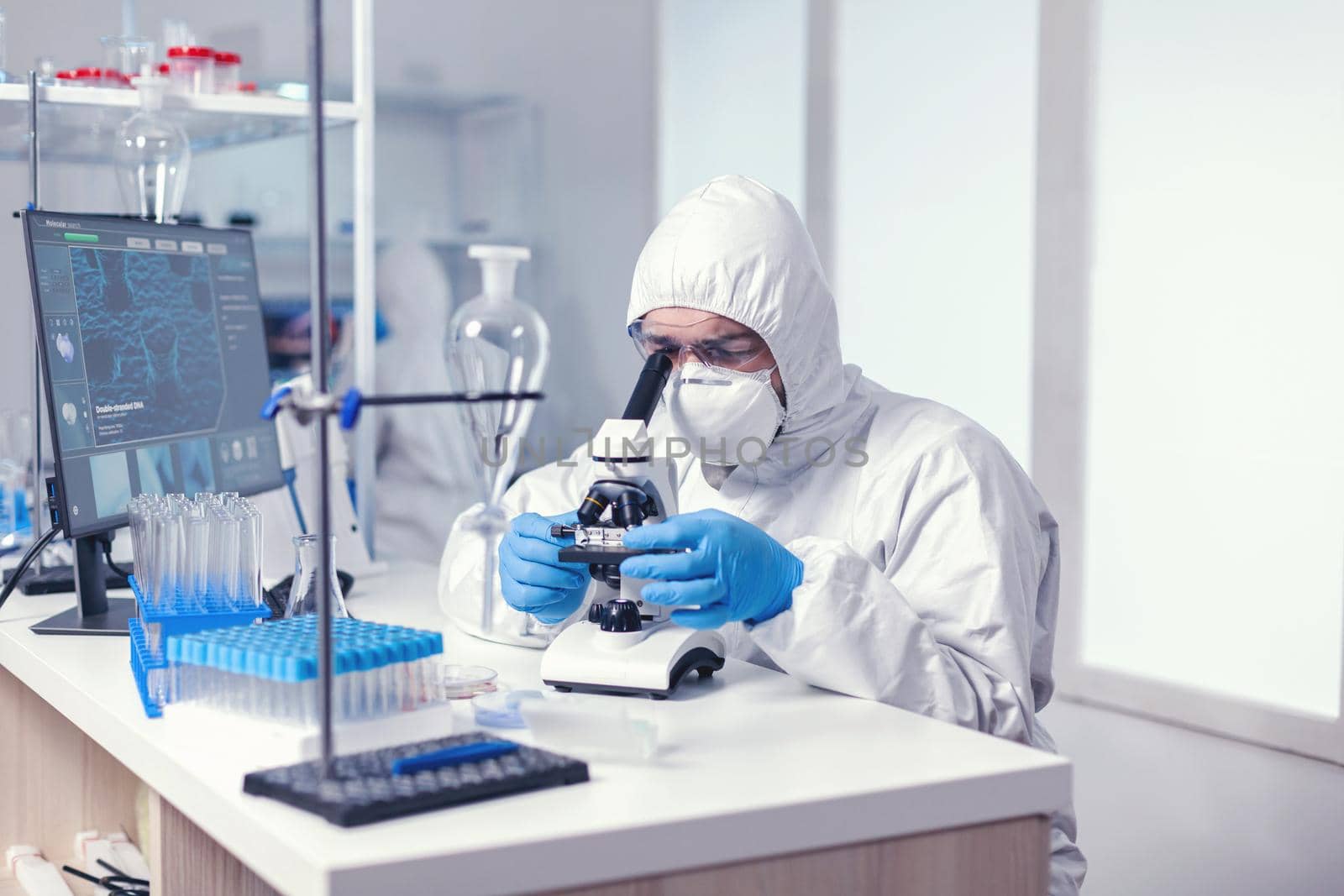 Biotechnology scientist in ppe suit researching cure for coronavirus using microscope in modern laboratory. Virolog in coverall during coronavirus outbreak conducting healthcare scientific analysis.