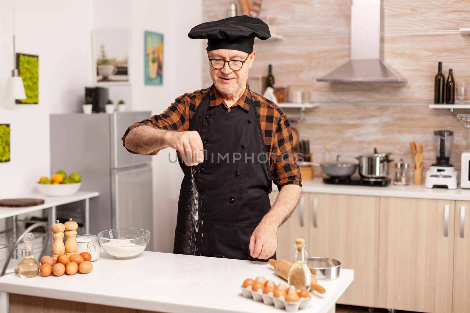 Elderly man preparing food spreading flour in home kitchen for food preparation. Retired senior chef with bonete and apron, in kitchen uniform sprinkling sieving sifting ingredients by hand.