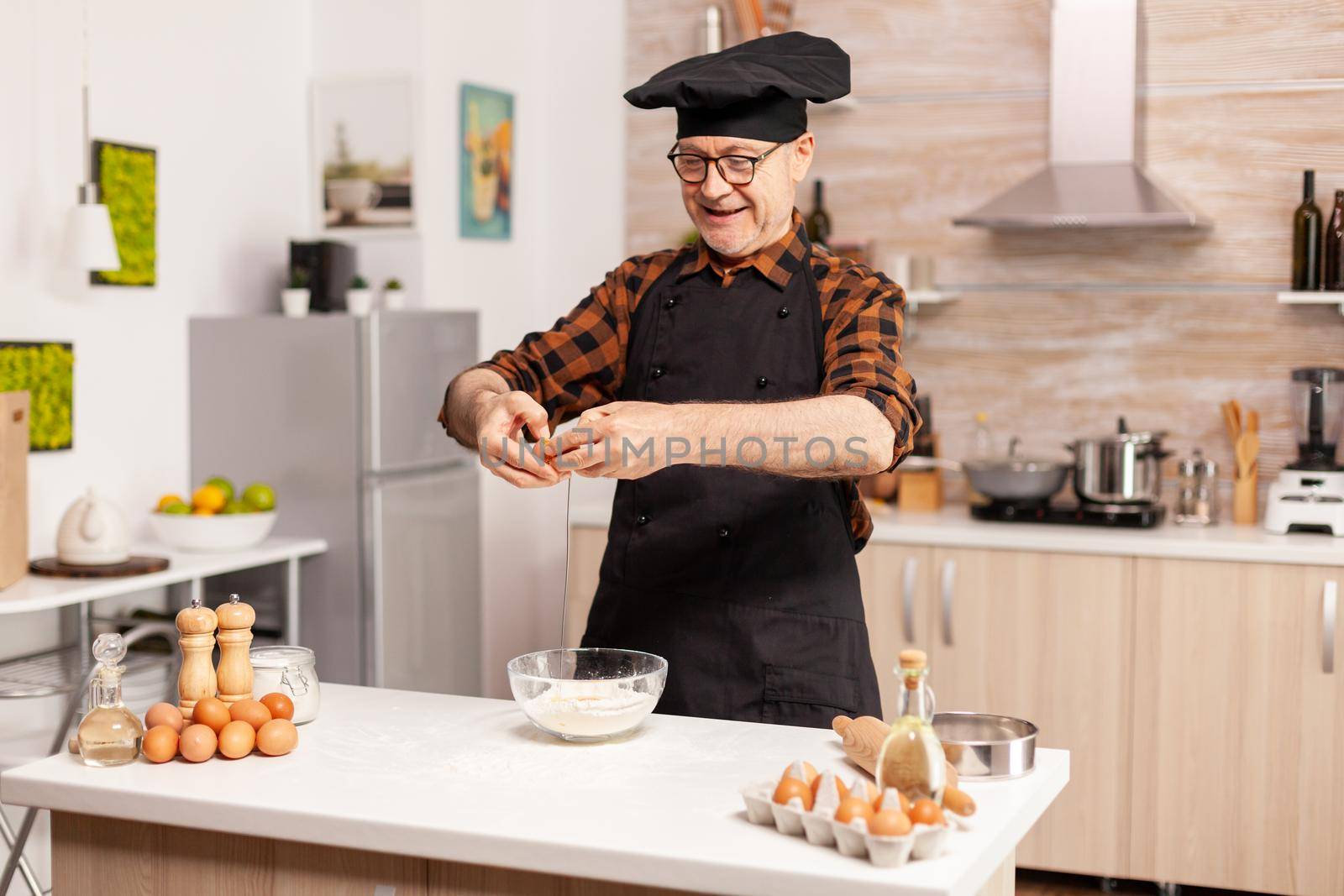 Happy senior man cracking eggs over wheat flour while preparing traditional recipe. Elderly pastry chef cracking egg on glass bowl for cake recipe in kitchen, mixing by hand, kneading