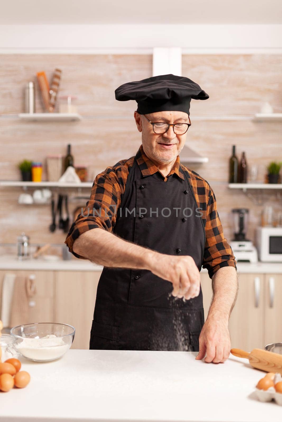 Cheerful sprinkles flour on the table to make dough for homemade bread. Retired senior chef with bonete and apron, in kitchen uniform sprinkling sieving sifting ingredients by hand.