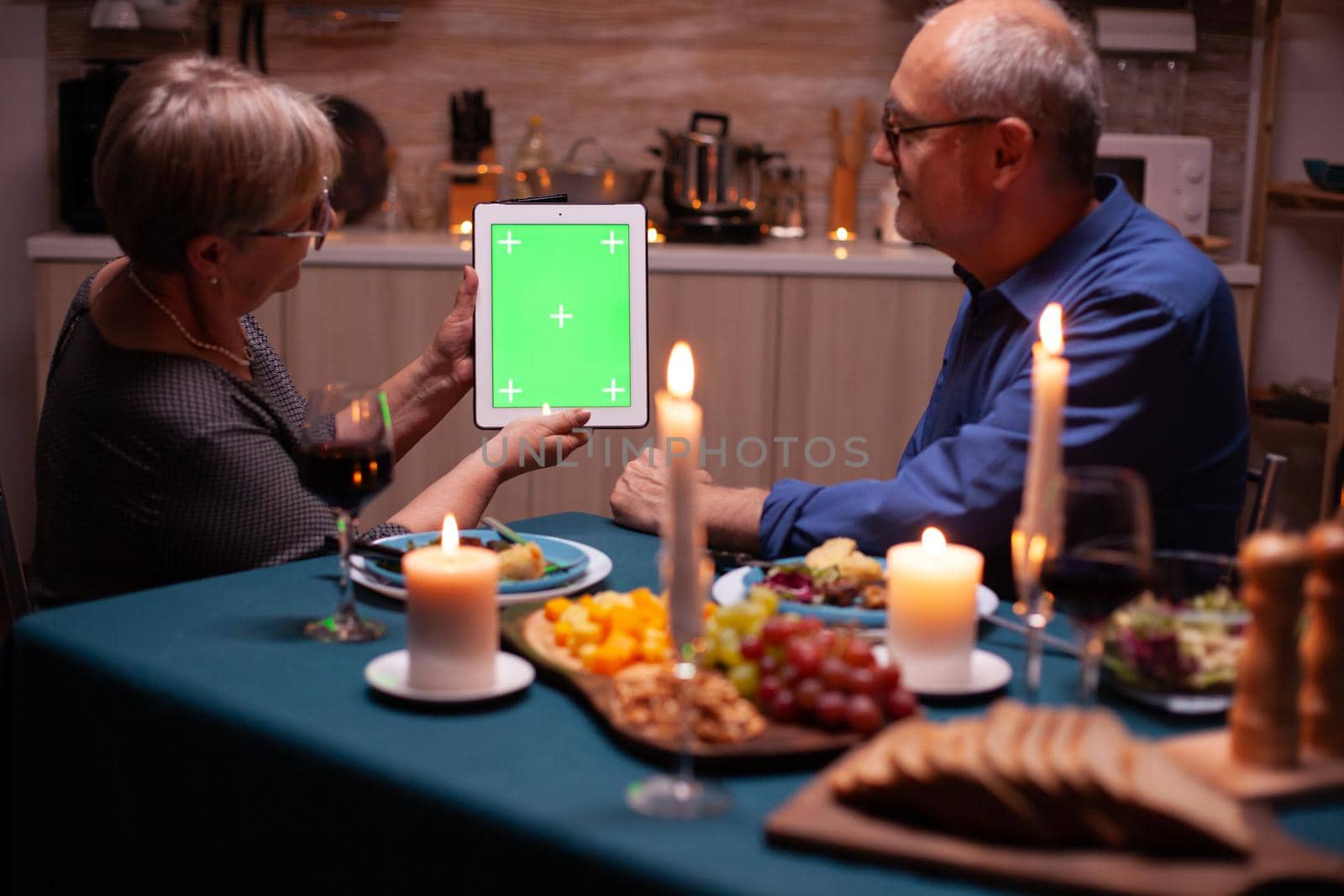 Old couple with green screen tablet pc in kitchen during festive dinner. Aged people looking at green screen template chroma key display sitting at the table in kitchen.