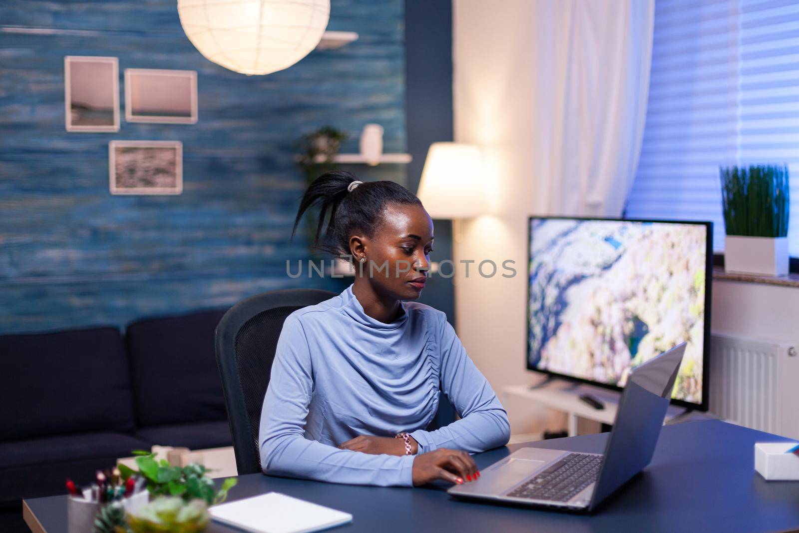 Dark skinned freelancer reading documentation on laptop computer in living room late at night. Black entrepreneur sitting in personal workplace writing on keyboard.