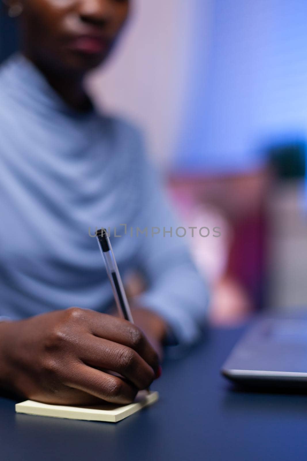 Close up of african woman taking notes sitting at desk in the evening for job. Black freelancer respecting deadline studying late at night.