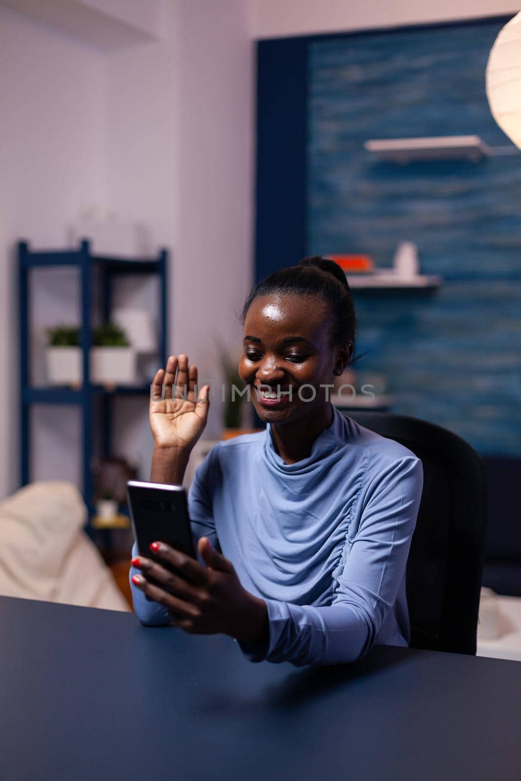 Dark skinnned woman saying hello in the course of video conference sitting at desk late at night in office. Black freelancer working with remotely team chatting virtual online conference.
