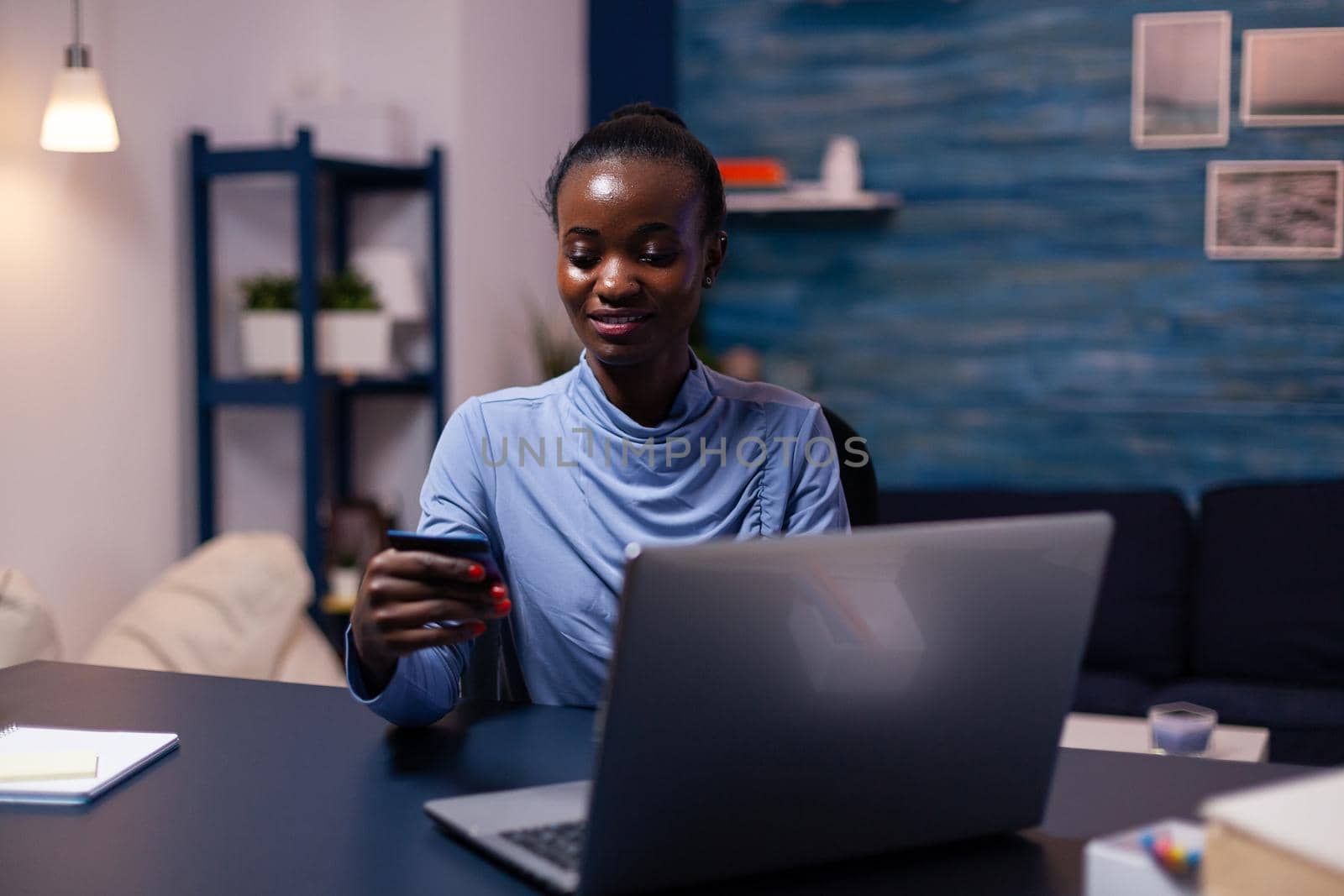 Dark skin young woman holding plastic credit card purchasing product from online shop. Employee making paymant transaction from home on digital notebook.