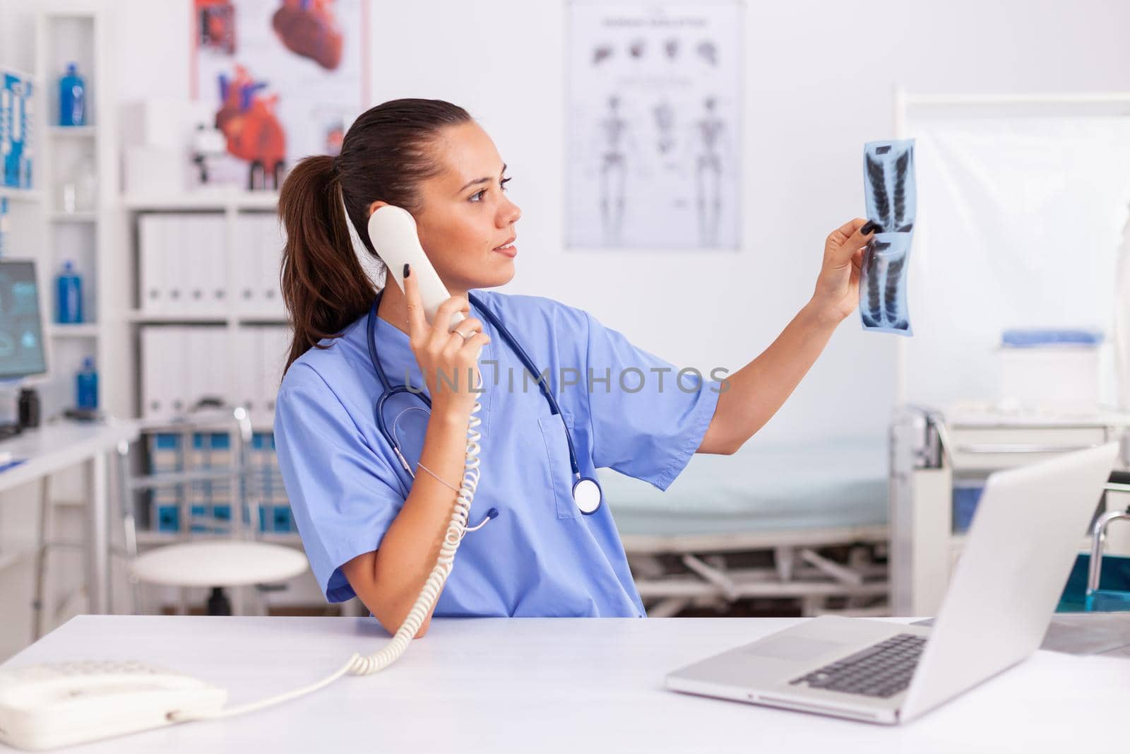 Medical nurse holding patient radiography in hospital office while talking with doctor on phone. Health care physician sitting at desk using computer in modern clinic looking at monitor.