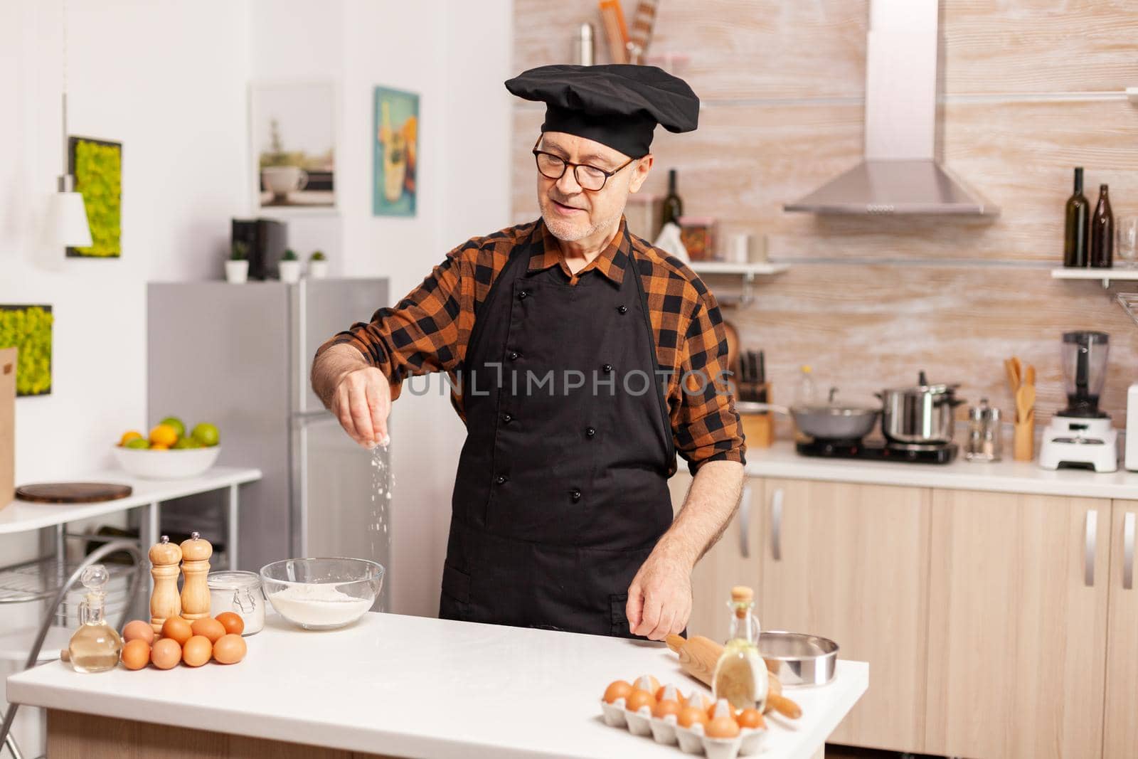 Senior baker in home kitchen putting wheat flour over wooden table. Retired senior chef with bonete and apron, in kitchen uniform sprinkling sieving sifting ingredients by hand.