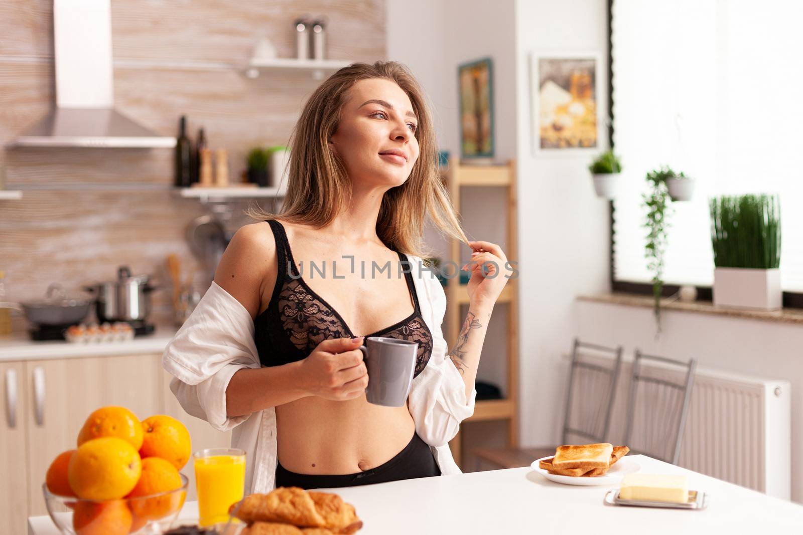 Woman wearing sexy bra during breakfast in home kitchen. Young attractive woman with tattoos in seductive underwear holding cup of tea relaxing in the kitchen smiling.