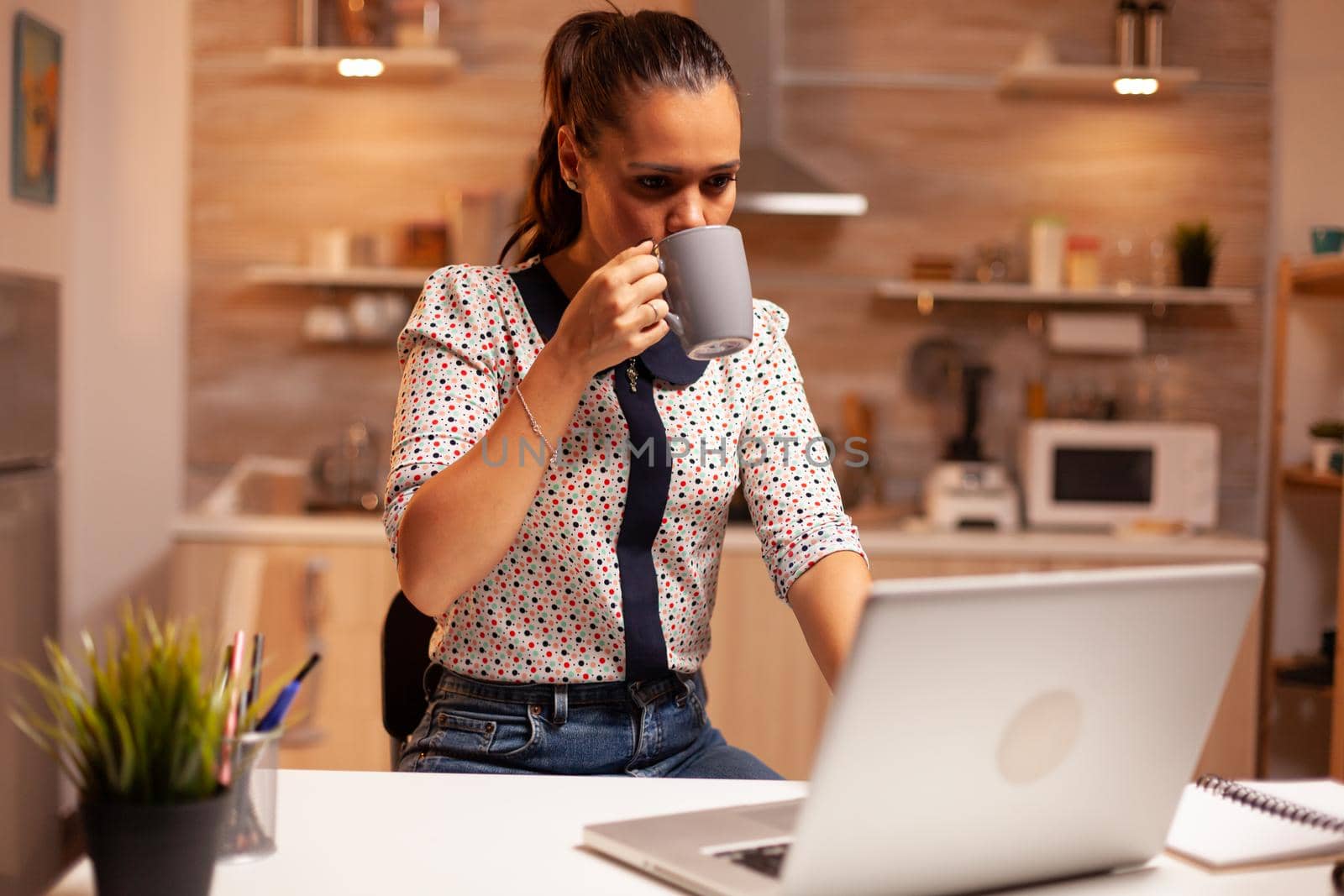 Woman taking a sip of coffee while working late at night on laptop in home kitchen for important project. Employee using modern technology at midnight doing overtime for job, business, busy, career.