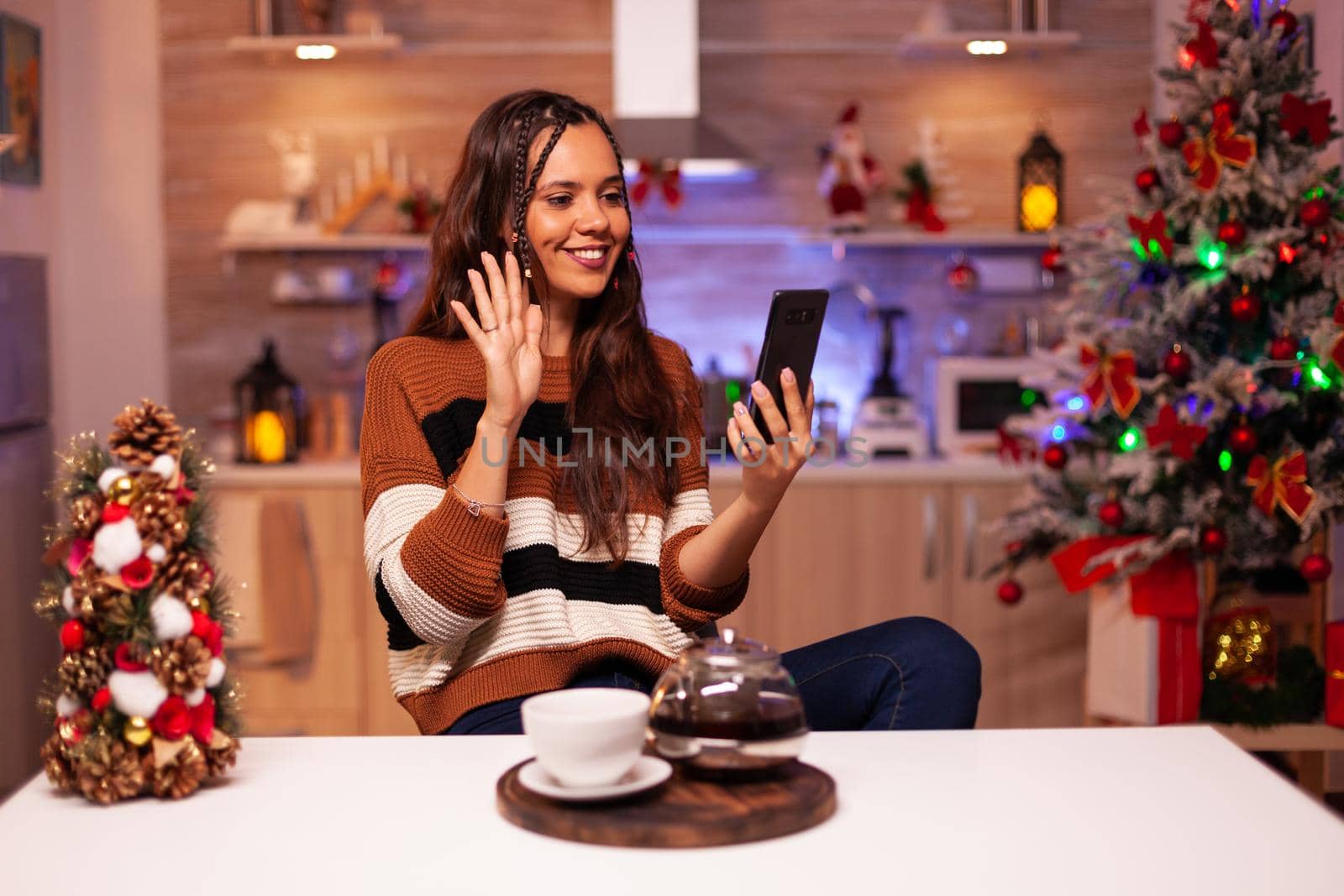Caucasian woman using video call technology on online internet, holding smartphone and cup of tea in winter decorated kitchen for holiday. Young smiling adult talking to family at home