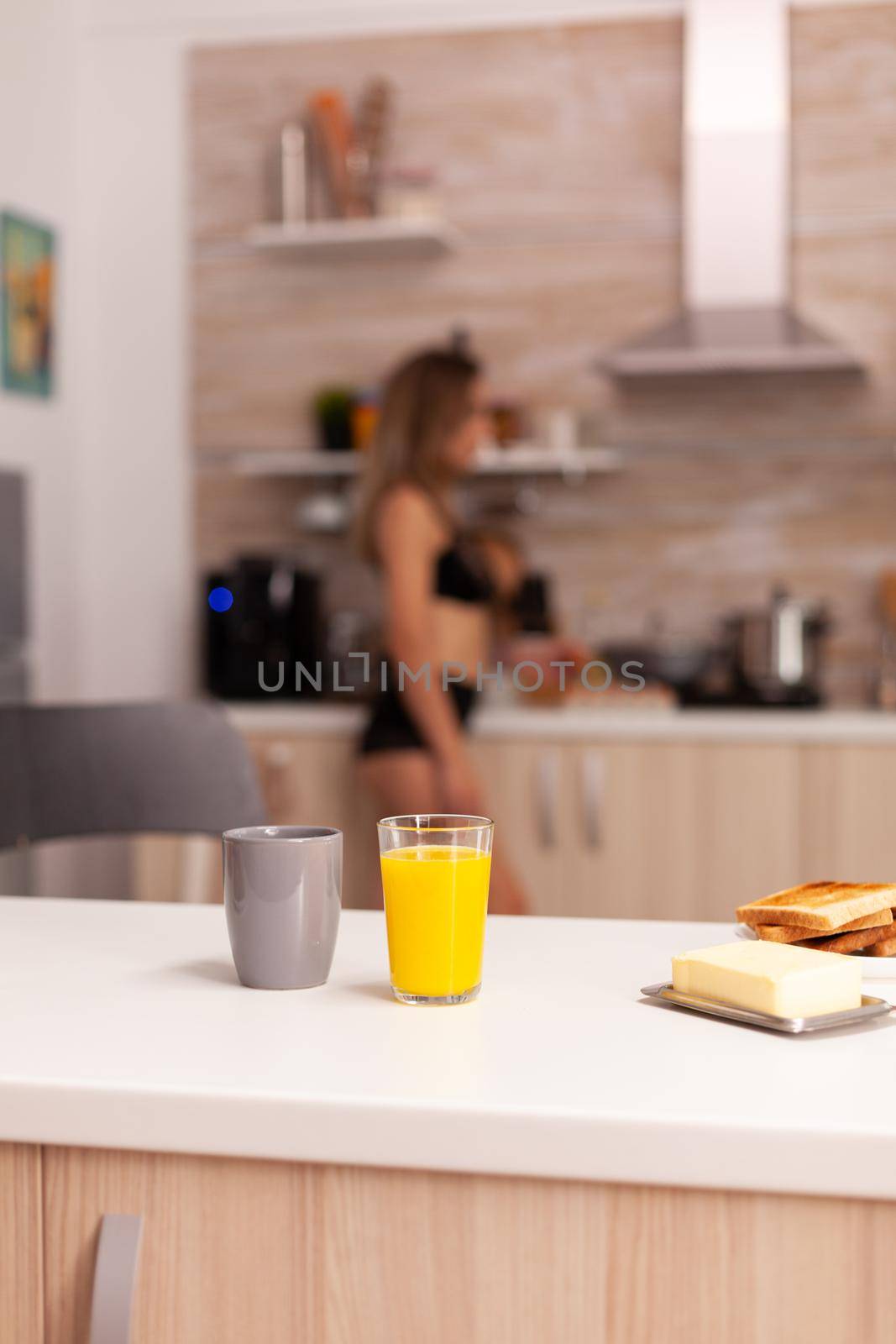 Cup of hot coffee on table during breakfast in home kitchen with carefree woman in black lingerie. Young sexy seductive blode lady with tattoos.