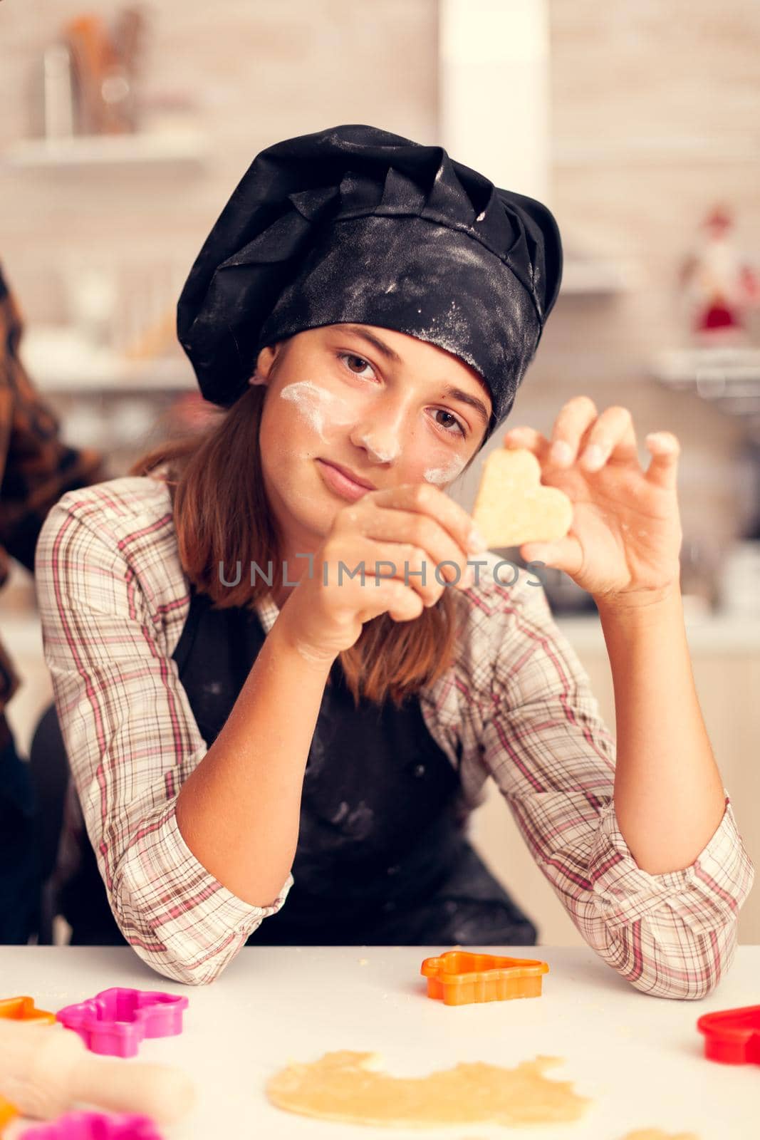 Childs hands with heart shaped pastry on christmas day wearing apron and bonette. Happy cheerful joyfull teenage girl helping senior woman preparing sweet cookies to celebrate winter holidays.