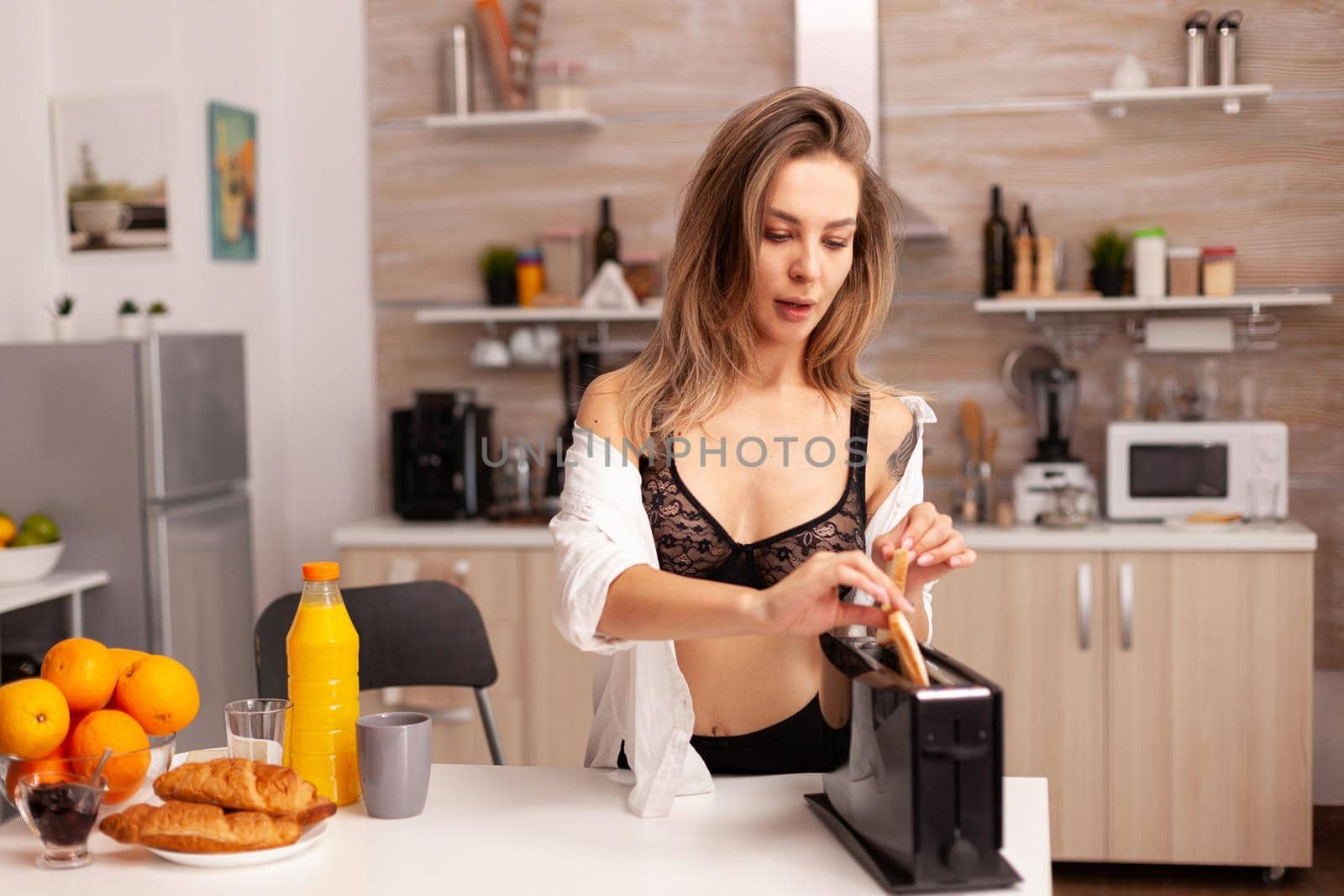 Sexy woman preparing roasted bread in home kitchen in lingerie. Young sexy seductive blode lady with tattoos drinking healthy, natural homemade orange juice, refreshing sunday morning.