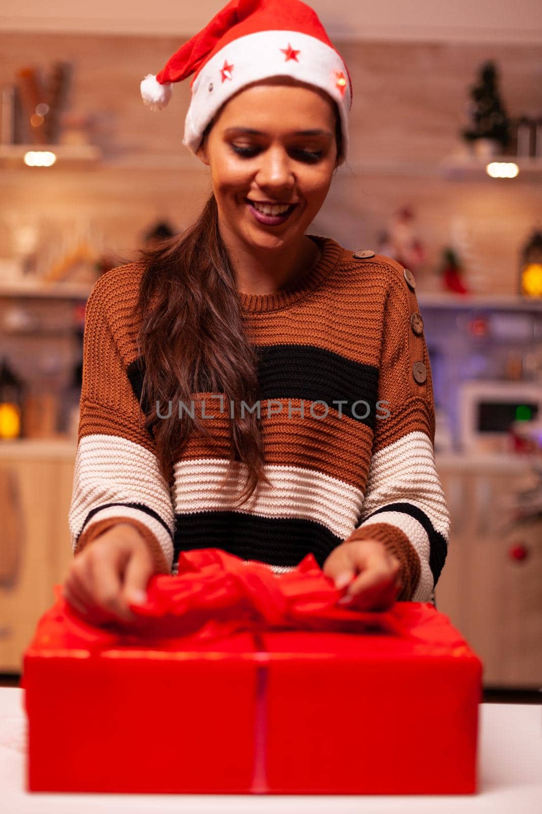 Traditional adult with santa hat using ribbon to tie bow on christmas gift box for friends at festive home. Young woman decorating present with wrapping paper for holiday celebration party