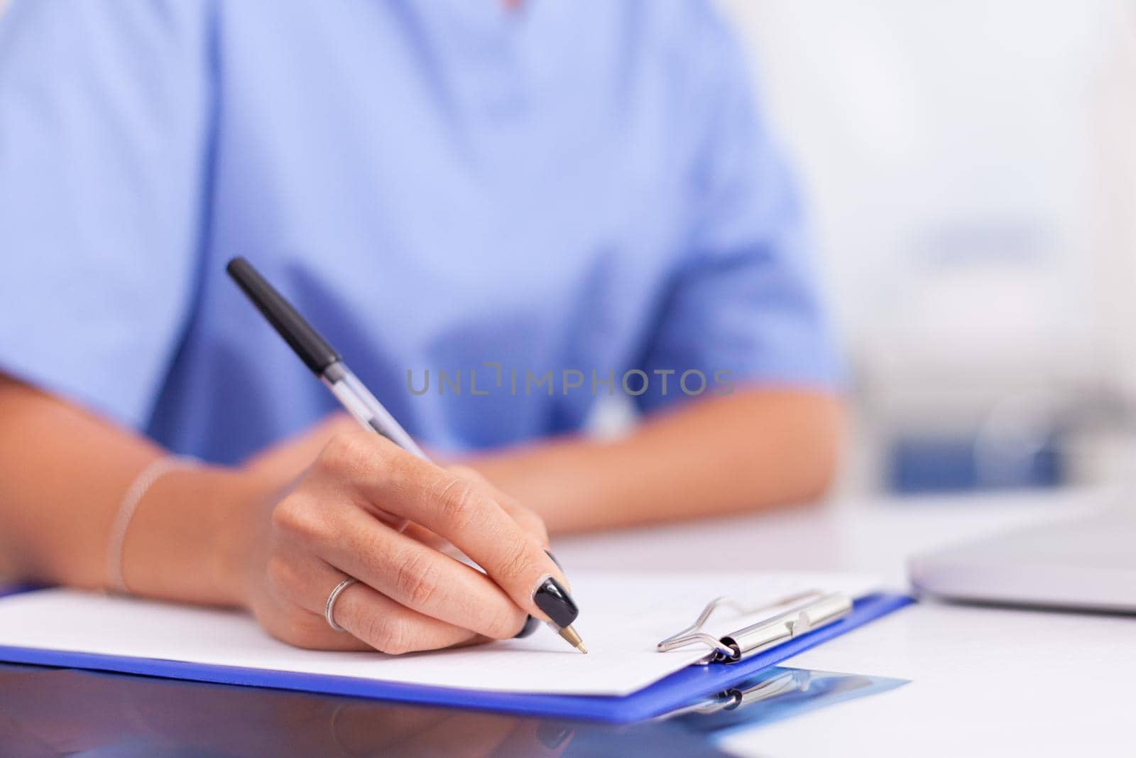 Close up of nurse writing patient diagnosis on clipboard in hospital office . Health care physician using computer in modern clinic looking at monitor, medicine, profession, scrubs.