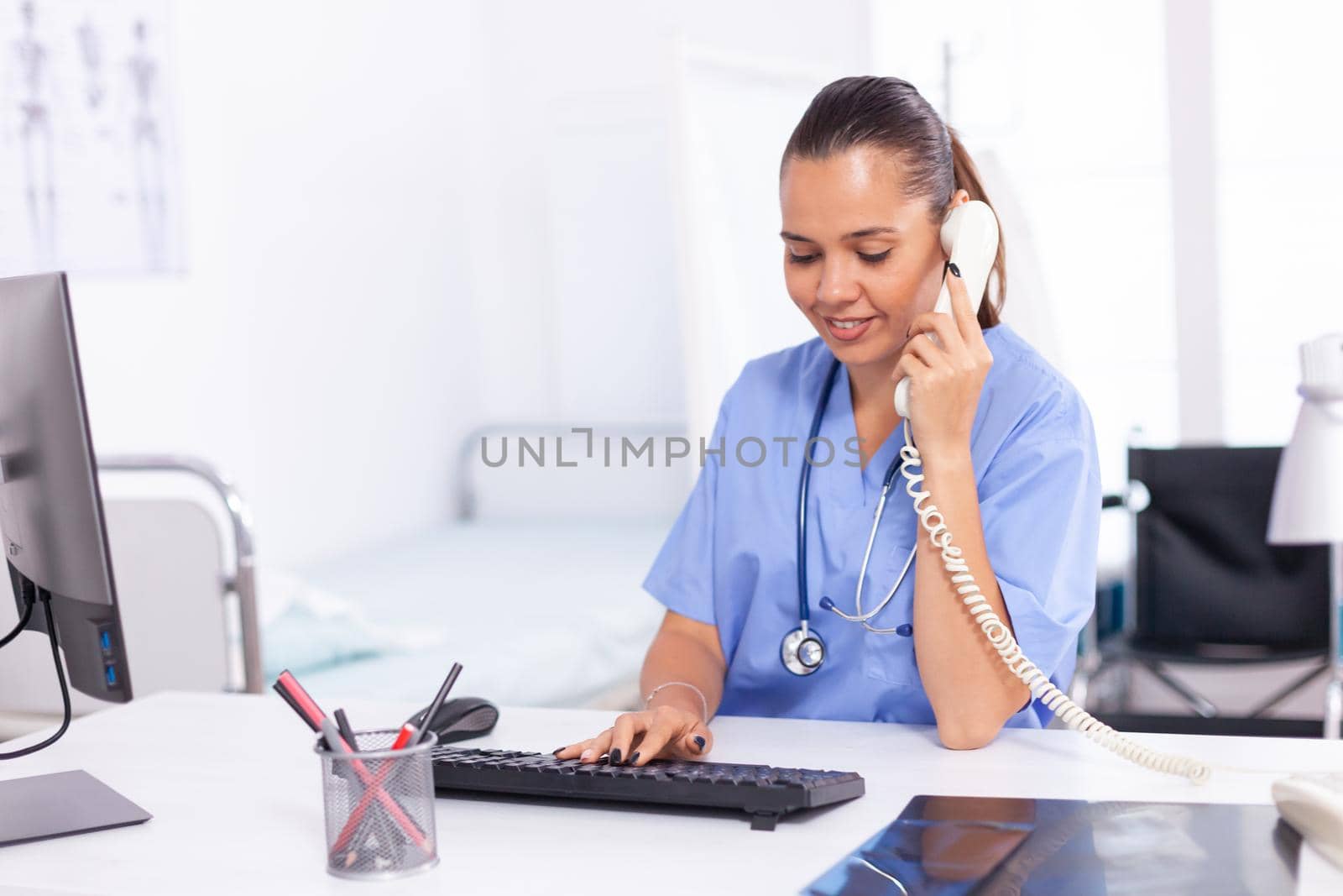 Nurse calling patient from hospital reception to talk about diagnosis. Health care physician sitting at desk using computer in modern clinic looking at monitor.