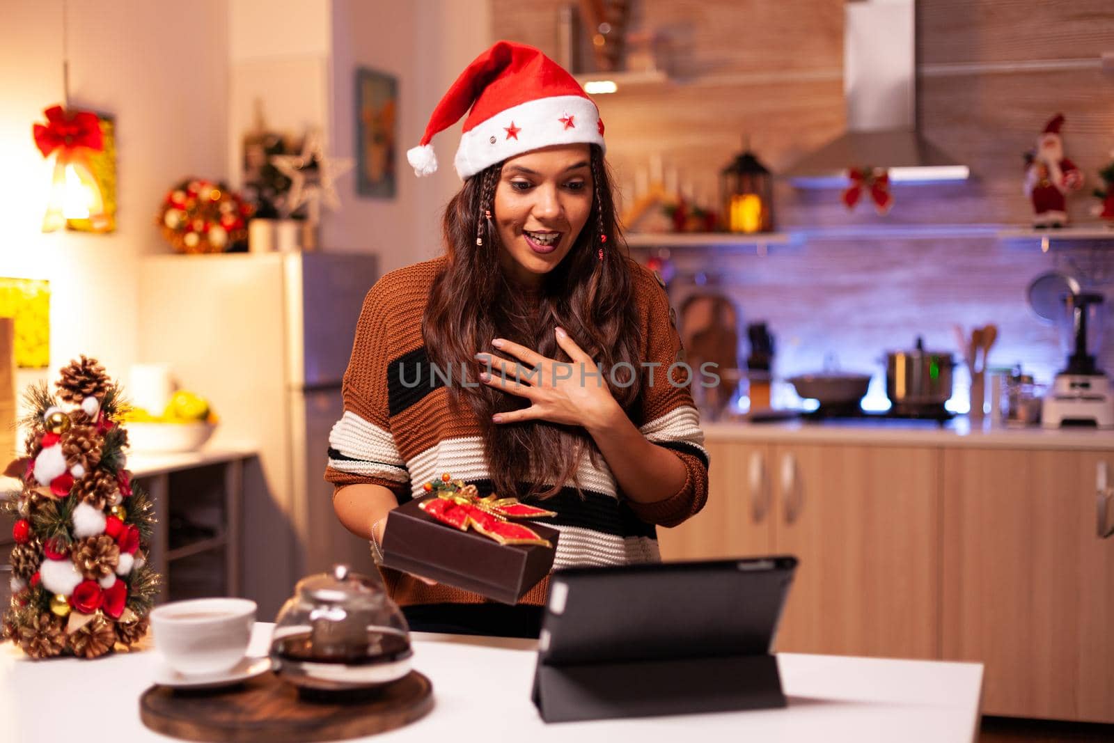 Young person using video call technology on tablet to give seasonal christmas present in festive decorated kitchen. Caucasian woman sharing winter joy with friends preparing for festivity