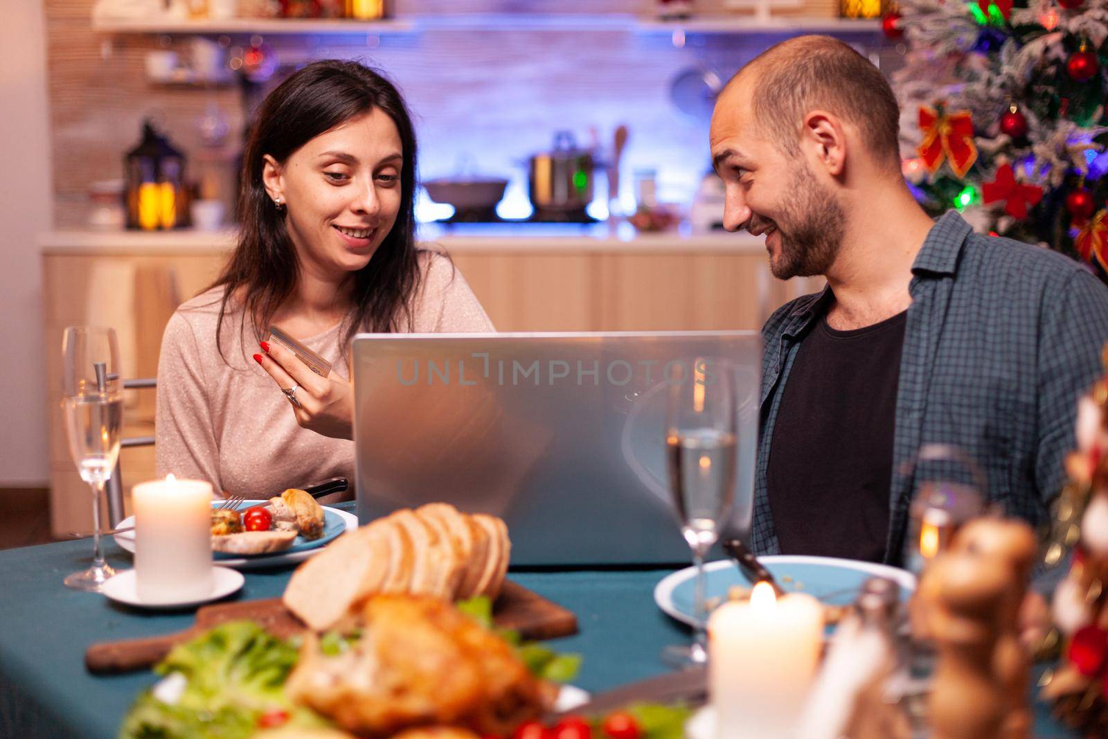 Happy family shopping xmas present gift using credit card doing online payment on laptop computer sitting at x-mas table in kitchen. Cheerful couple celebrating christmas holiday