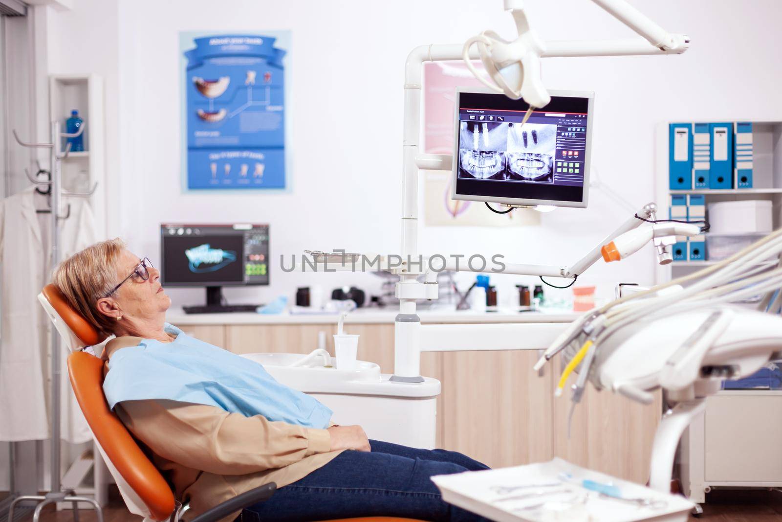 Senior woman waiting stomatolog sitting on orange chair for consultation. Elderly patient during medical examination with dentist in dental office with orange equipment.