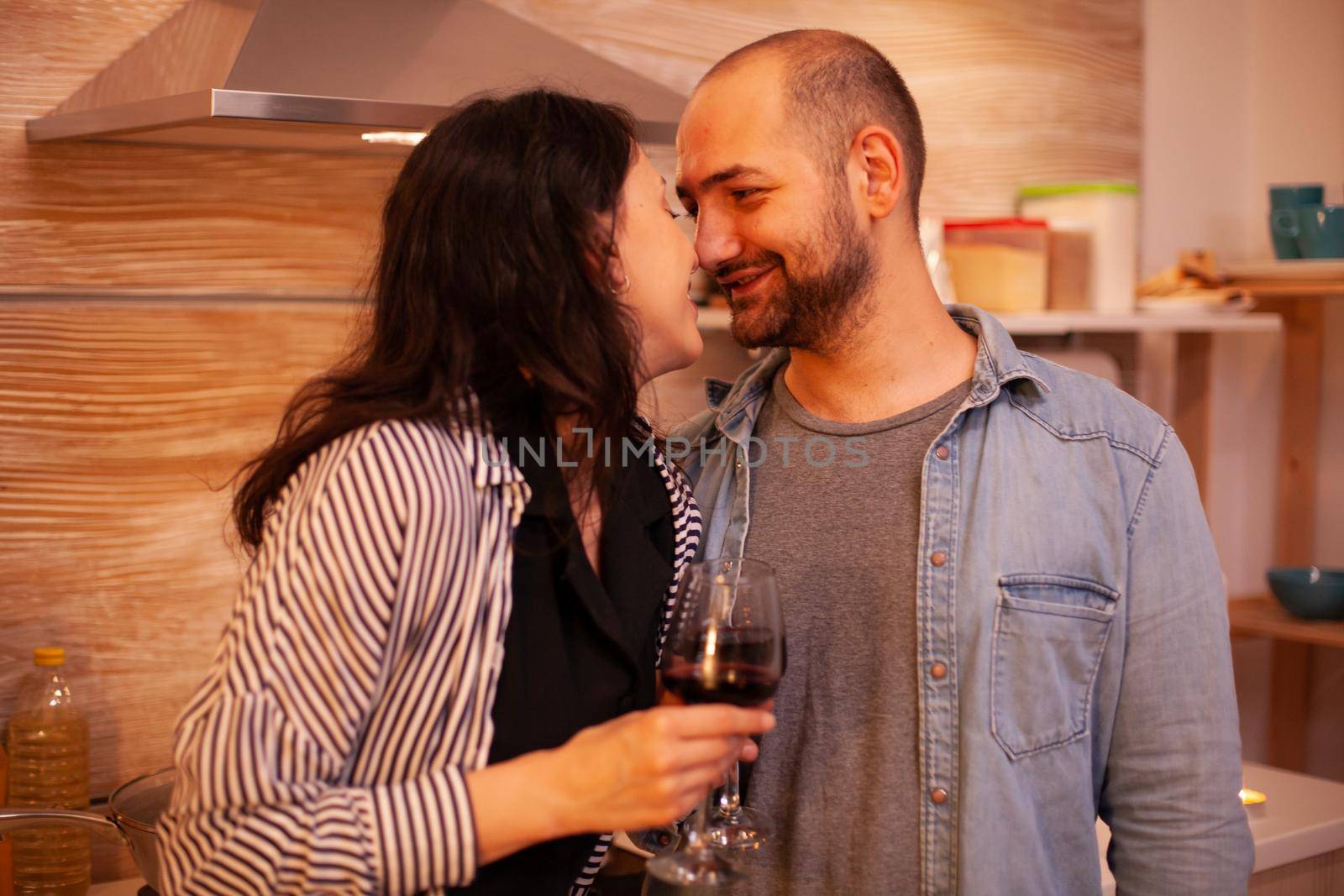 Couple touching noses and having romance date in kitchen. Adult couple at home, drinking red wine, talking, smiling, enjoying the meal in dining room.