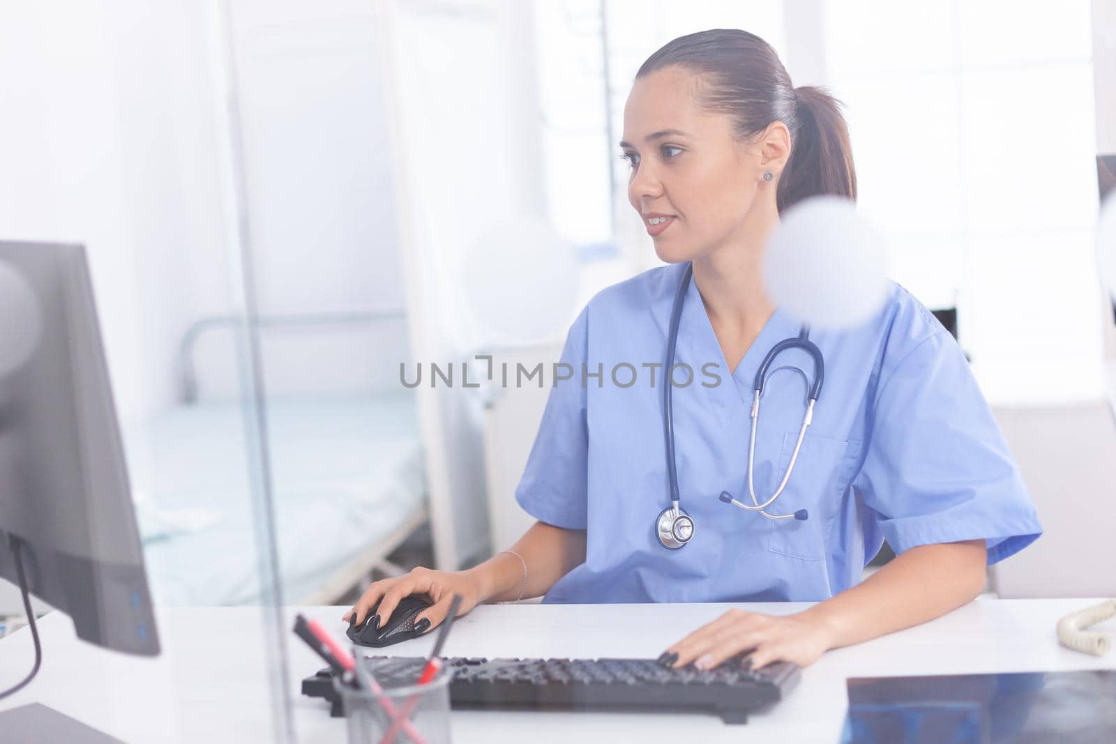 Medical practitioner using computer in hospital office. Health care physician using computer in modern clinic looking at monitor, medicine, profession, scrubs.