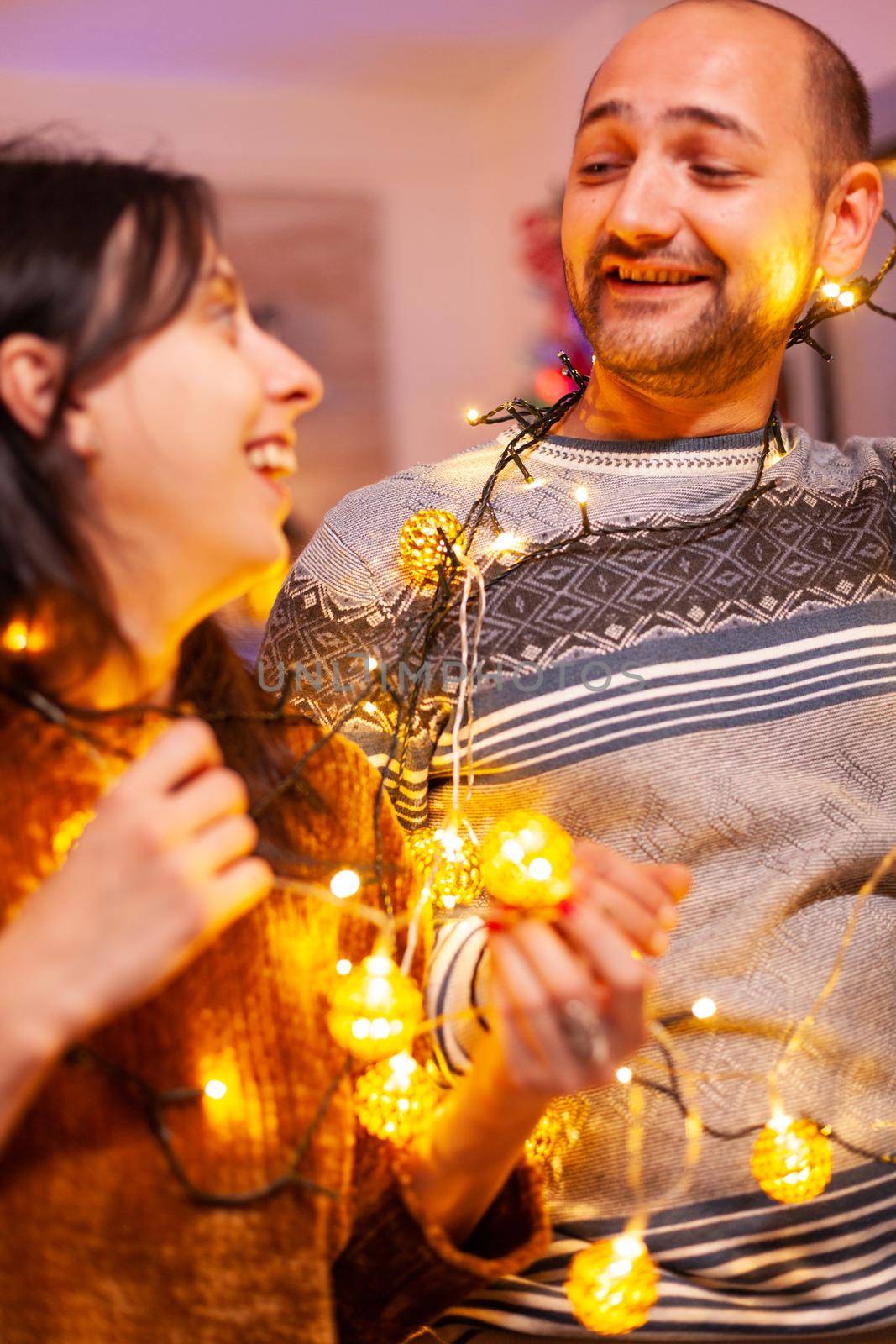 Family stuck in christmas tree lights in xmas decorated kitchen celebrating christmas holiday. Happy couple making funny while enjoying spending winter season together. New-year festive tradition