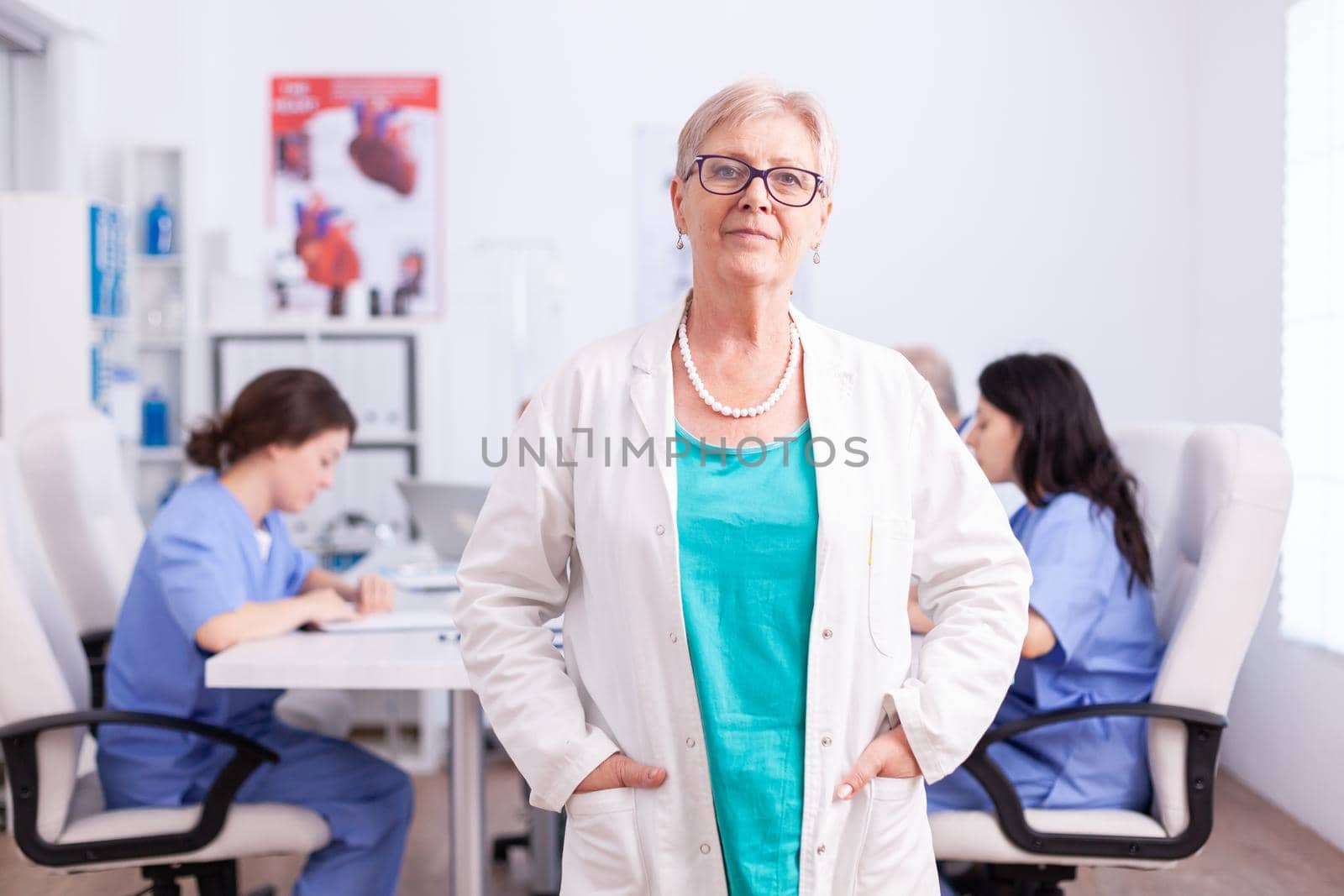 Portrait of senior doctor smiling at camera in hospital conference room with medical staff in the background.