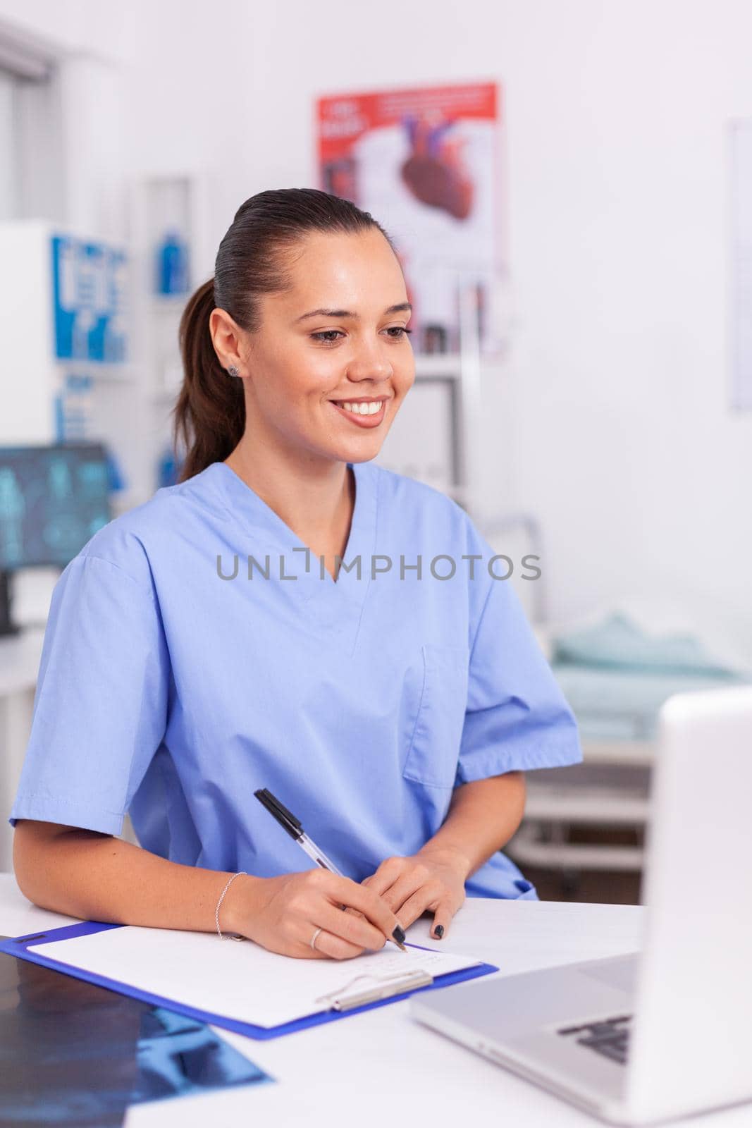 Smiling medical nurse writing notes on clipboard sitting at desk in hospital. Health care physician using computer in modern clinic looking at monitor, medicine, profession, scrubs.