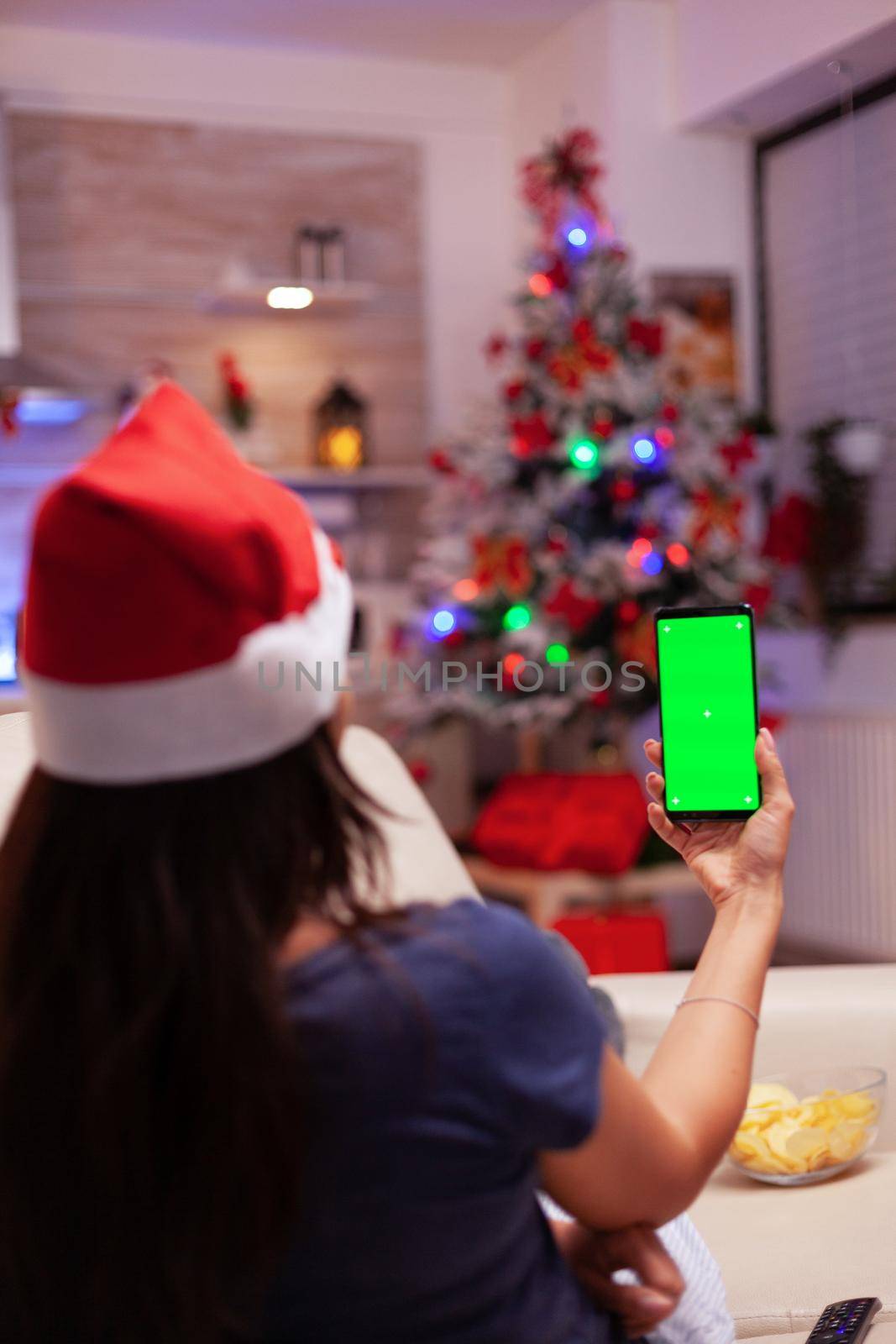 Woman holding green screen mock up chroma key smartphone with isolated display during christmastime in xmas decorated kitchen. Girl enjoying winter season celebrating christmas holiday