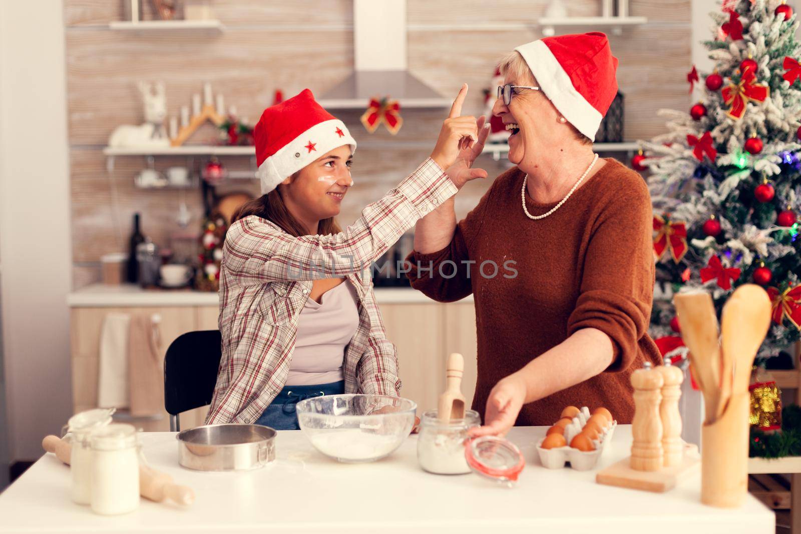 Grandmother and niece on christmas day having fun making cakes. Happy cheerful joyfull teenage girl helping senior woman preparing sweet cookies to celebrate winter holidays wearing santa hat.