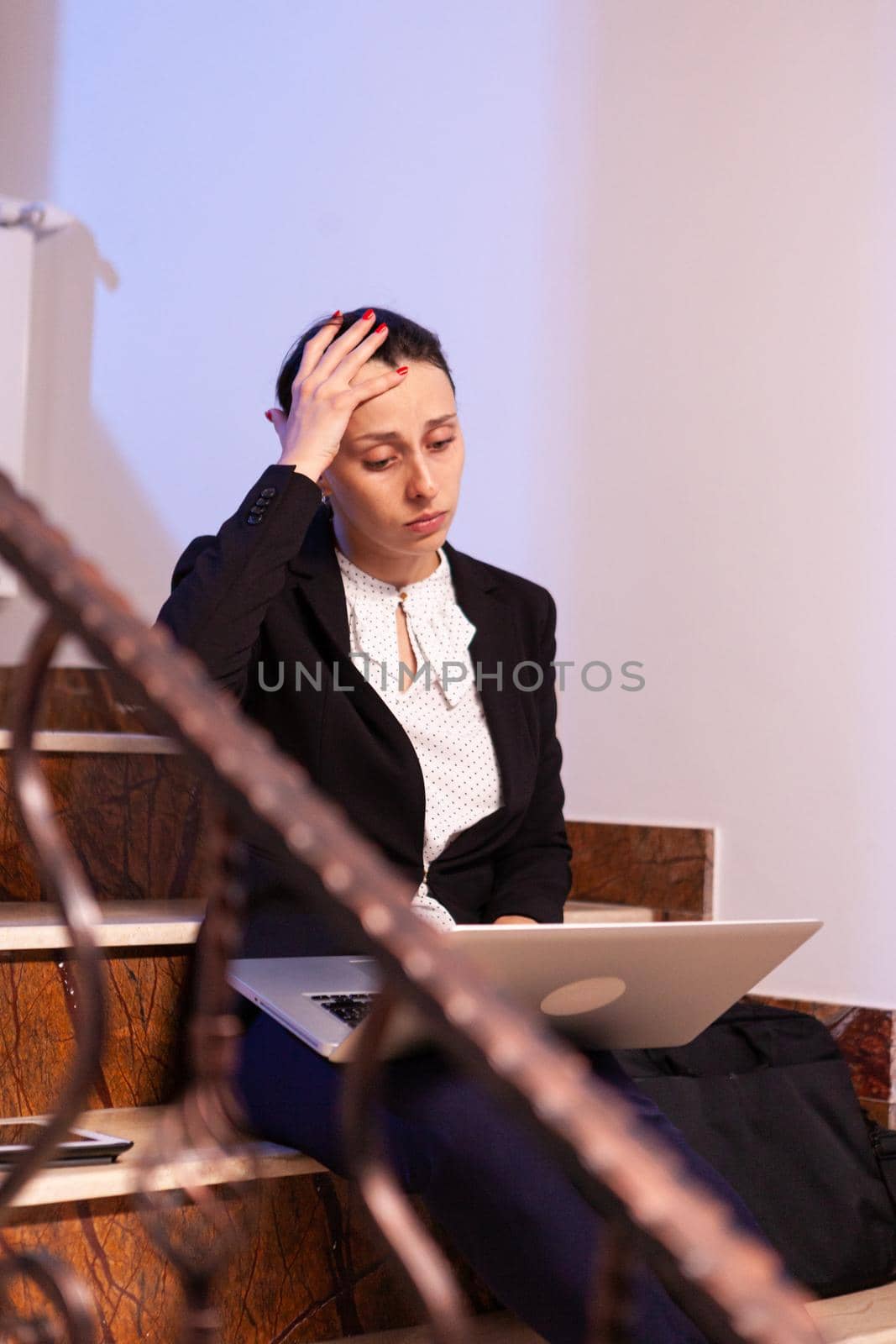 Overworked tired businesswoman with headache working on difficult project deadline. Serious entrepreneur working on job project sitting on staircase of business building at night hours for job.