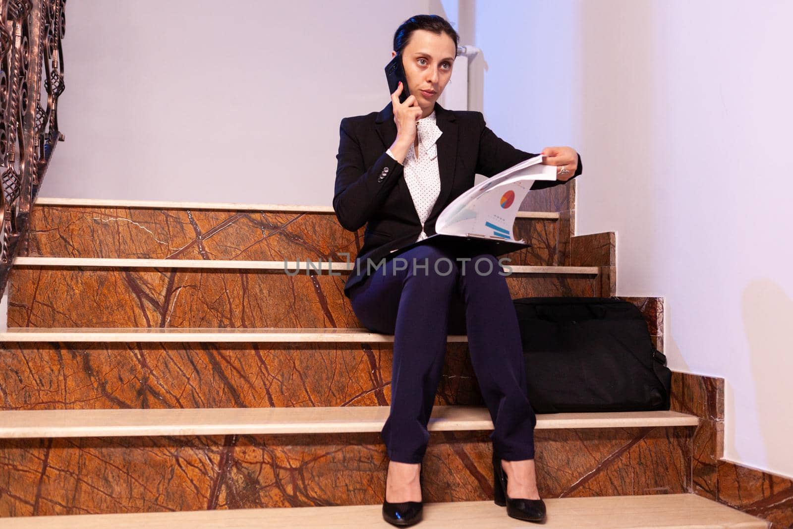 Overworked tired businesswoman reading project deadline during phone call with coworker. Serious entrepreneur working on job project sitting on staircase of business building at night hours for job.