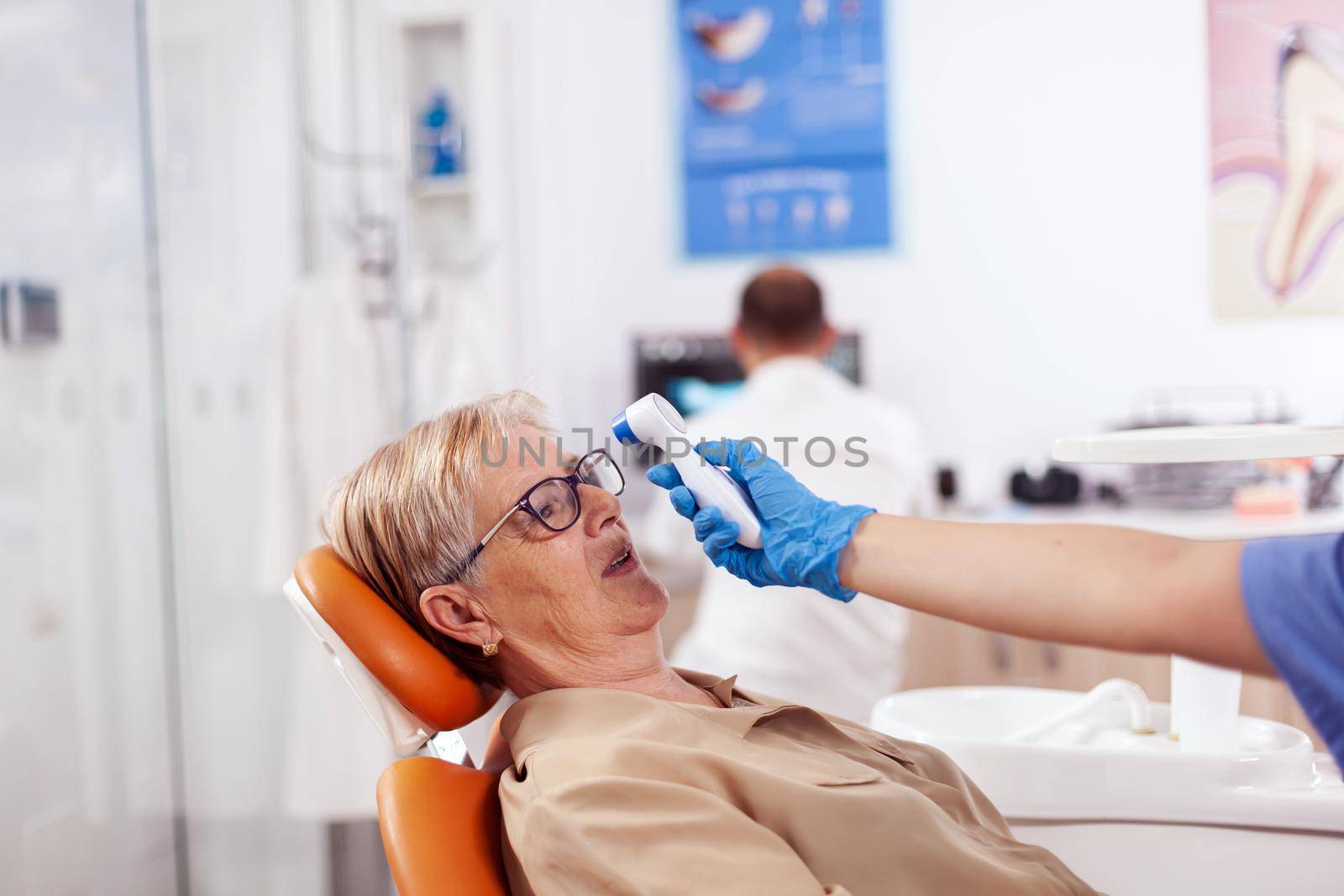 Dentist assistant holding digital body temperature indicator in front of patient forhead sitting on chair. Medical specialist in dental clinic taking patient temperature using digital device.