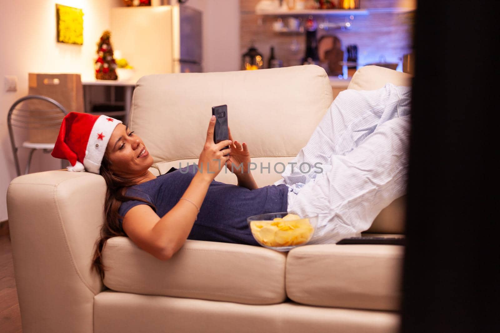 Girl messaging with friend using smartphone resting on couch in xmas decorated kitchen. Woman browsing on social media, texting during christmas holiday enjoying winter season