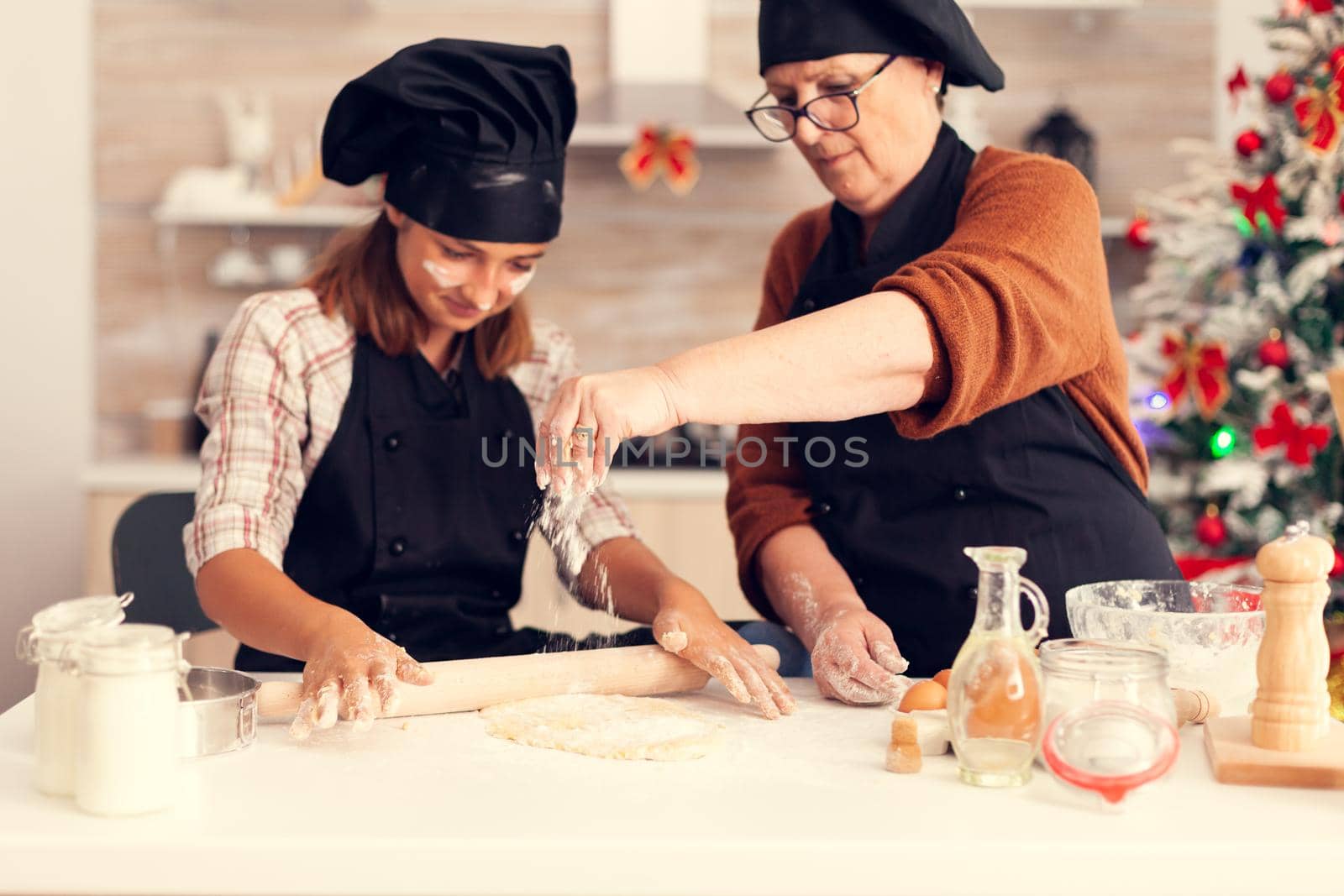 Grandmother wearing apron on christmas day spreading flour Happy cheerful joyfull teenage girl helping senior woman preparing sweet cookies to celebrate winter holidays.