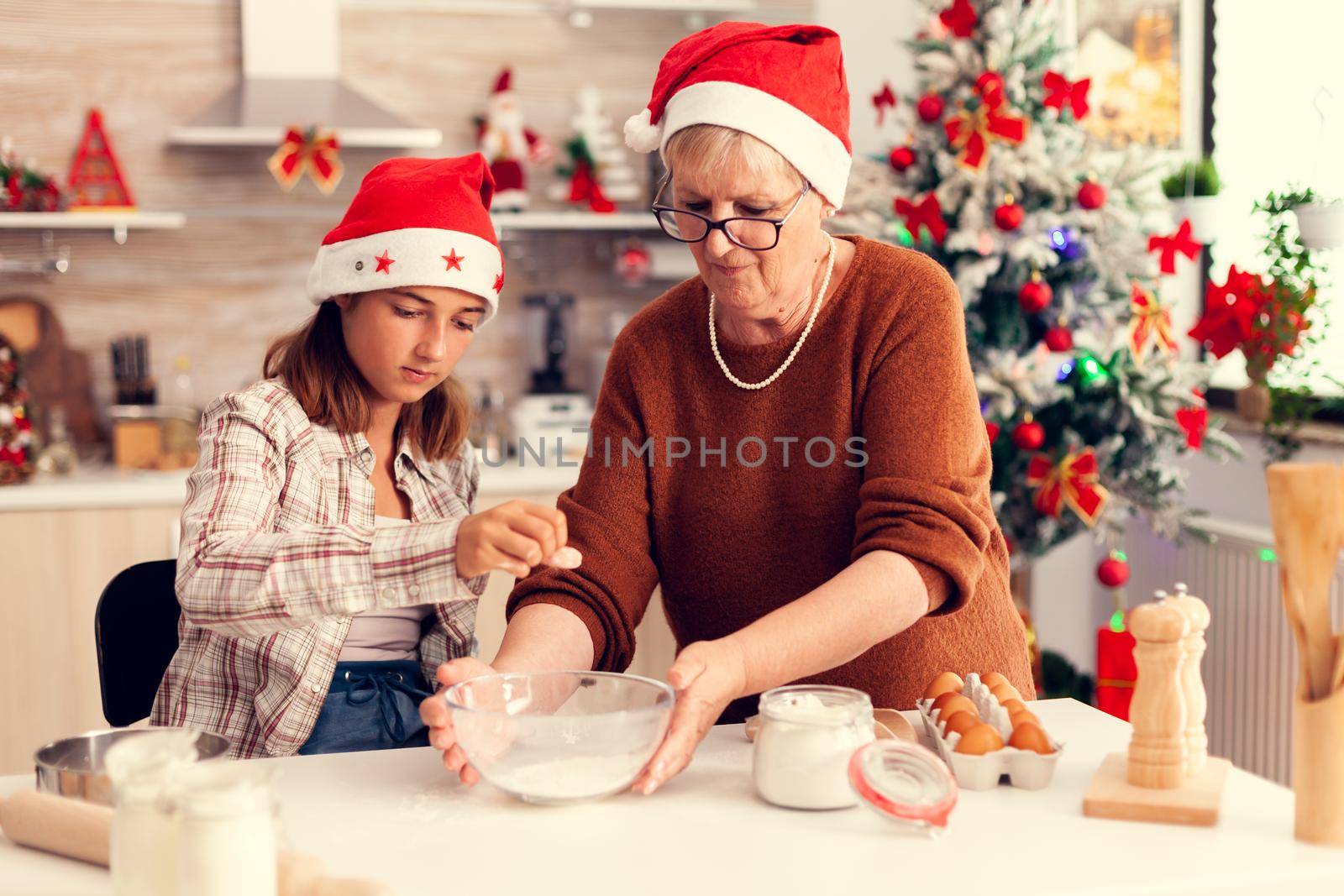 Loving grandmother on christmas day making cookies with niece. Happy cheerful joyfull teenage girl helping senior woman preparing sweet cookies to celebrate winter holidays wearing santa hat.