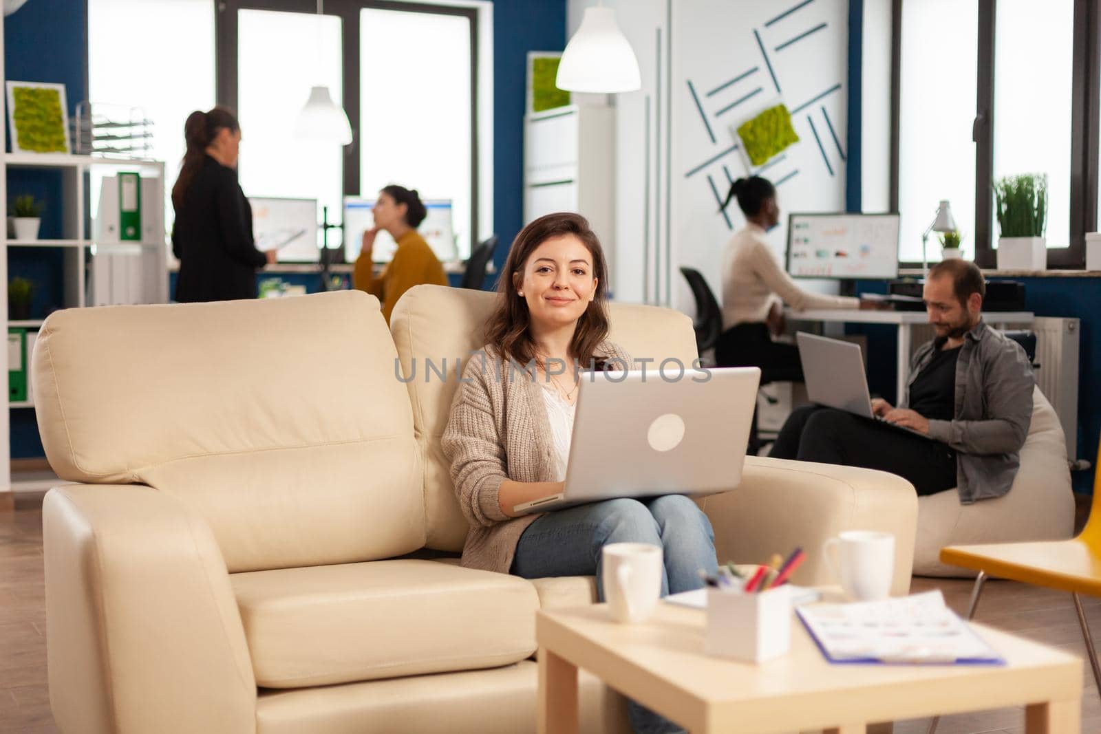 Happy manager woman sitting on sofa in front of camera smiling holding laptop while diverse colleagues working in background. Multiethnic coworkers analysing startup financial reports in modern office