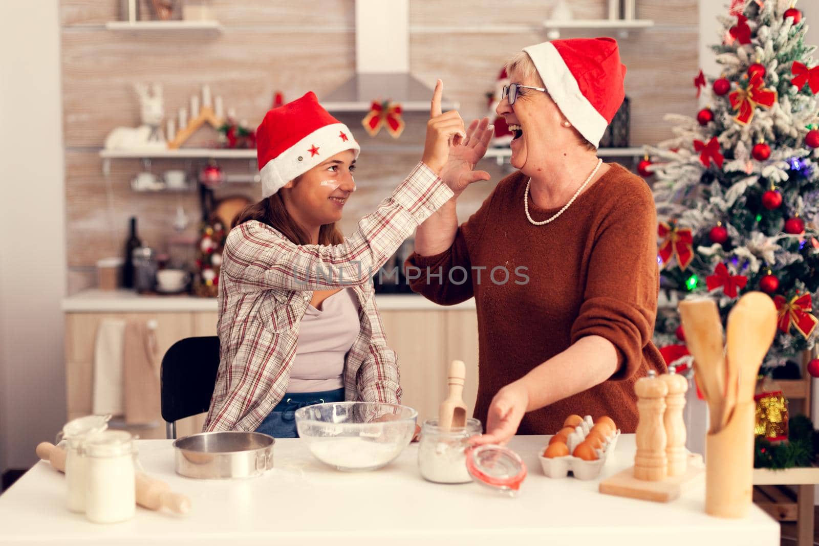 Niece and granddmother on christmas day enjoying making cakes and cookies. Happy cheerful joyfull teenage girl helping senior woman preparing sweet biscuits to celebrate winter holidays wearing santa hat.