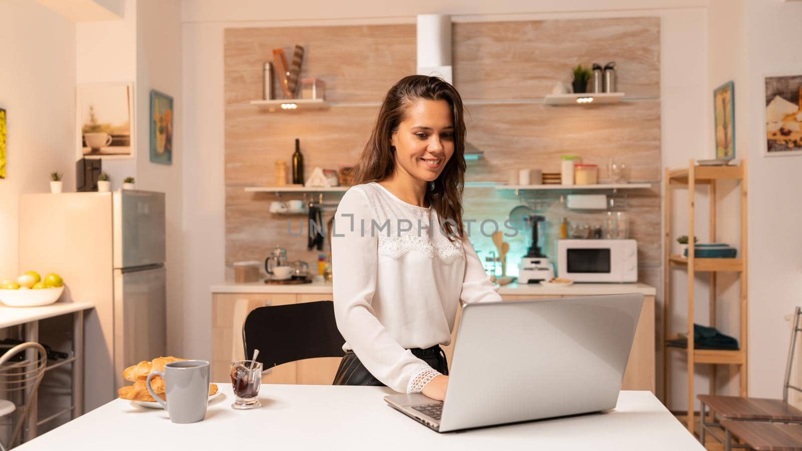 Busy woman in home kitchen using laptop to work on corporate project for job. Concentrated entrepreneur in home kitchen using notebook during late hours in the evening.