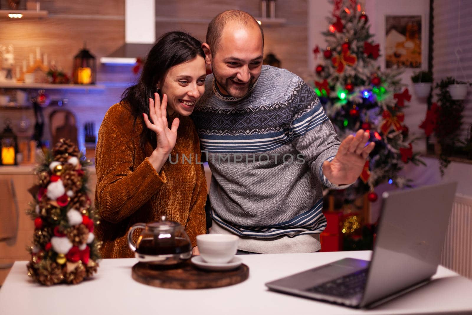 Happy family greeting friends during online videocall meeting on laptop computer celebrating christmas holiday together. Cheerful couple enjoying spending winter season in xmas decorated kitchen