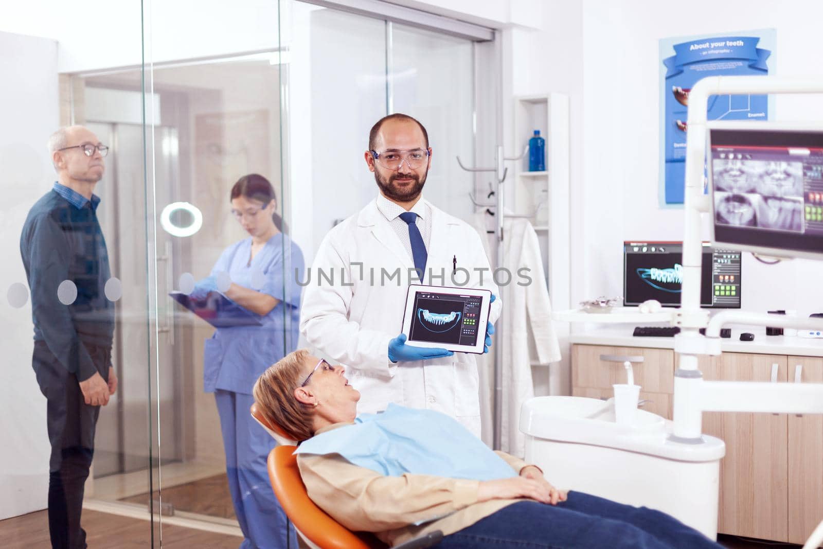 Stomatolog holding x-ray of senior woman sitting on orange chair in dentist cabinet. Medical teeth care taker holding patient radiography on tablet pc near patient standing up.