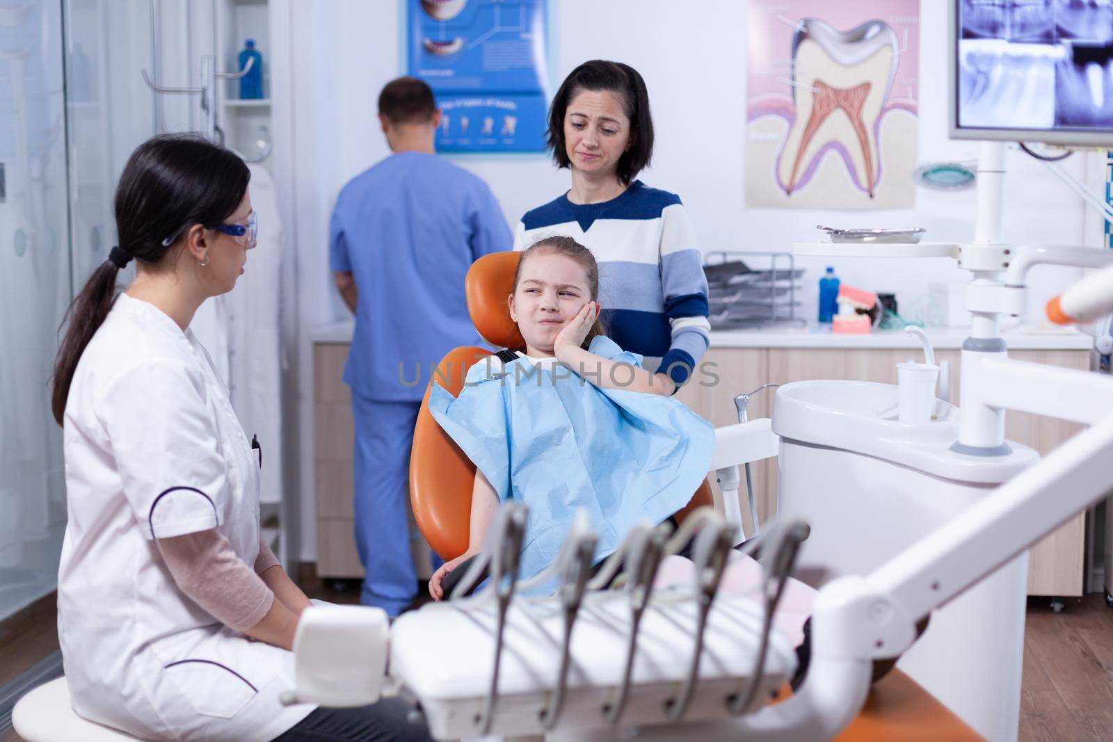 Little girl with painful expression showing dentist where her tooth hurts during dental check up. Child with her mother during teeth check up with stomatolog sitting on chair.