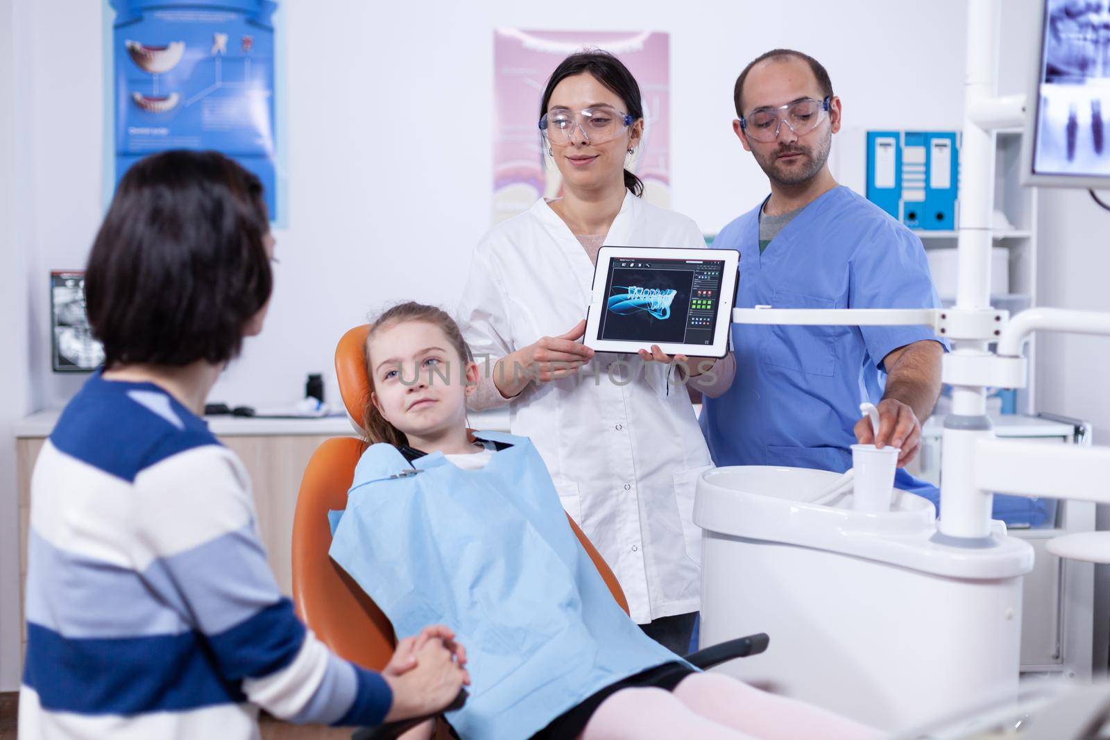 Dentist and patient commenting cavity treatment in tablet application after consultation. Stomatologist explaining teeth diagnosis to mother of child in health clinic holding x-ray.